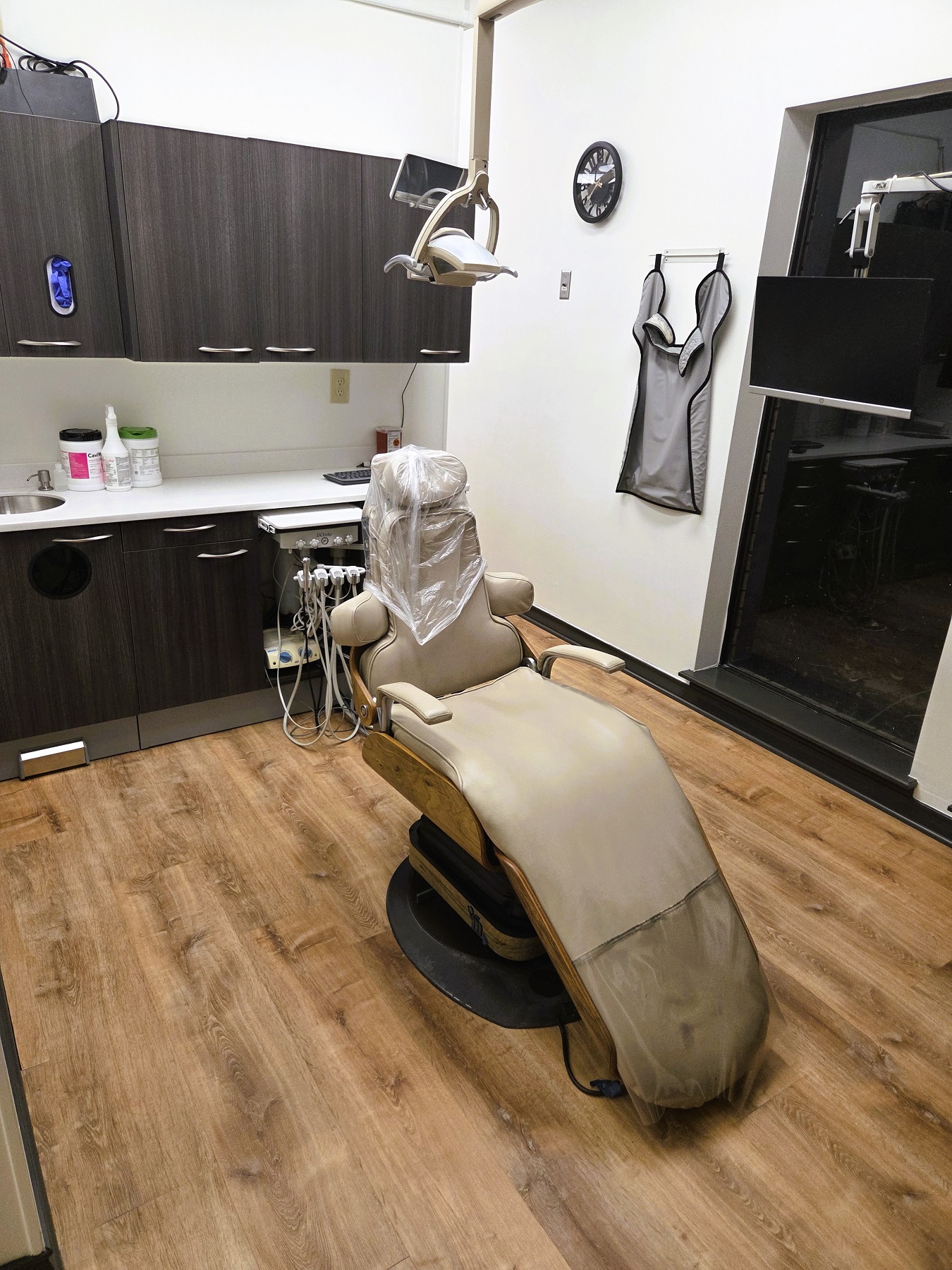 Light walls with dark stained baseboards and chair rail and medium stained wood flooring. A cushioned dental procedural chair anchors the room with an overhead dental light. A cabinet with sink, shelves hold lots of equipment, tools, and supplies.