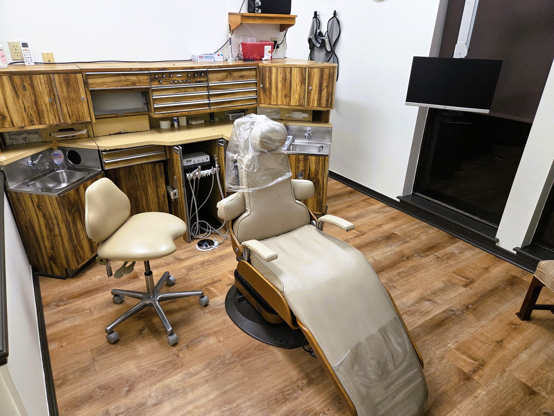 Light walls with dark stained baseboards and chair rail and medium stained wood flooring. A cushioned dental procedural chair anchors the room with an overhead dental light. A cabinet with sink, shelves hold lots of equipment, tools, and supplies.