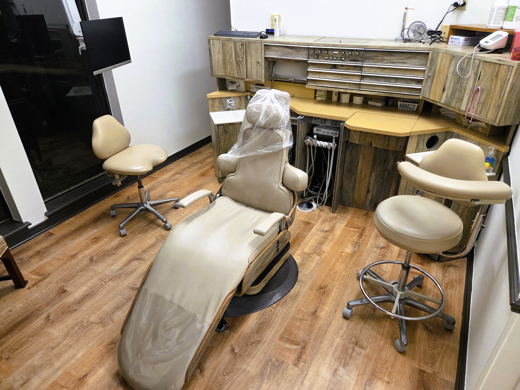 Light walls with dark stained baseboards and chair rail and medium stained wood flooring. A cushioned dental procedural chair anchors the room with an overhead dental light. A cabinet with sink, shelves hold lots of equipment, tools, and supplies.