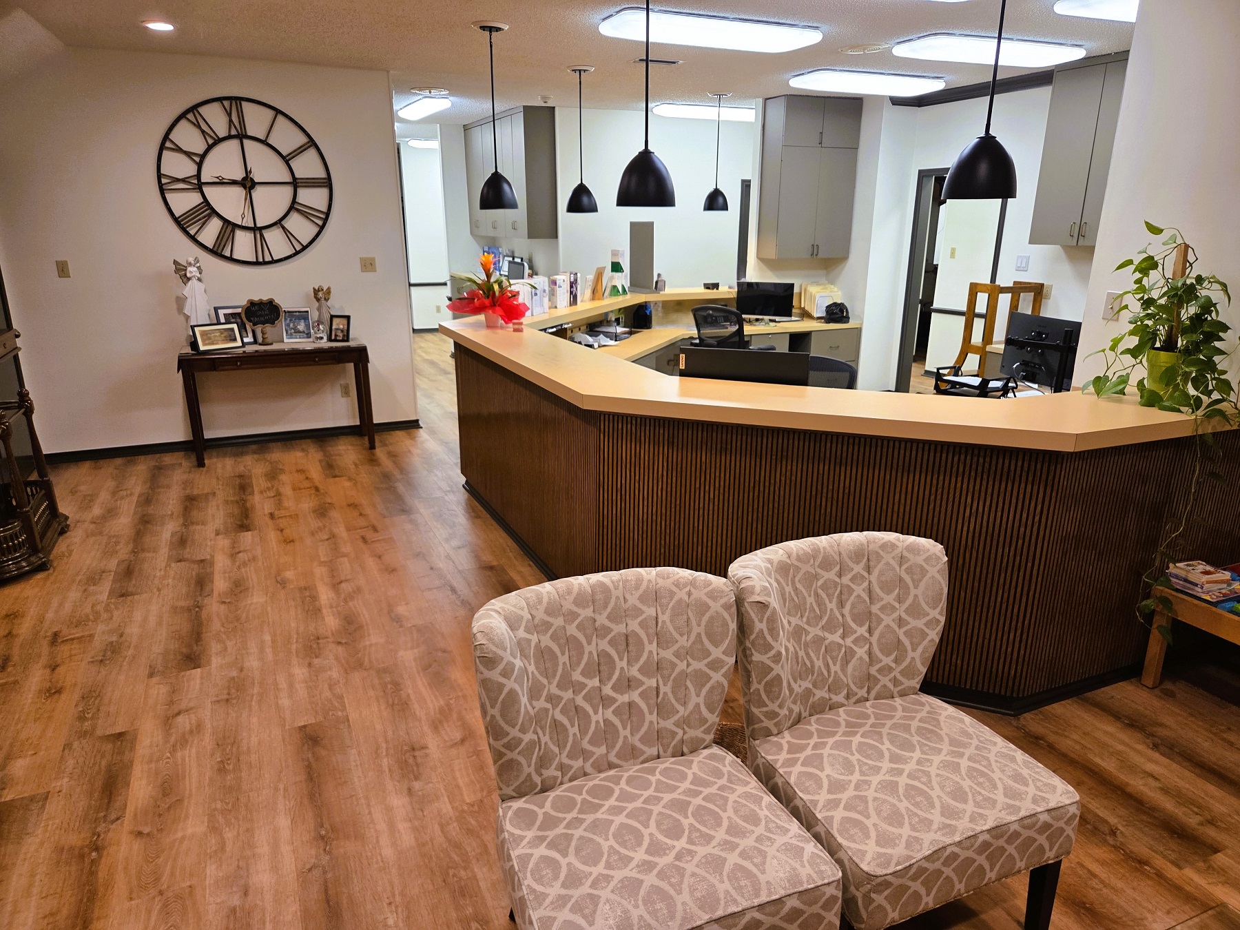 Light walls with dark stained wood baseboards and medium stained wood flooring. Darker stained wood curved reception desk with a curved yellow counter top and dark pendant lights hanging over it. A large clock hangs on the wall over a side table with magazines. A pair of light patterned cushioned chairs.