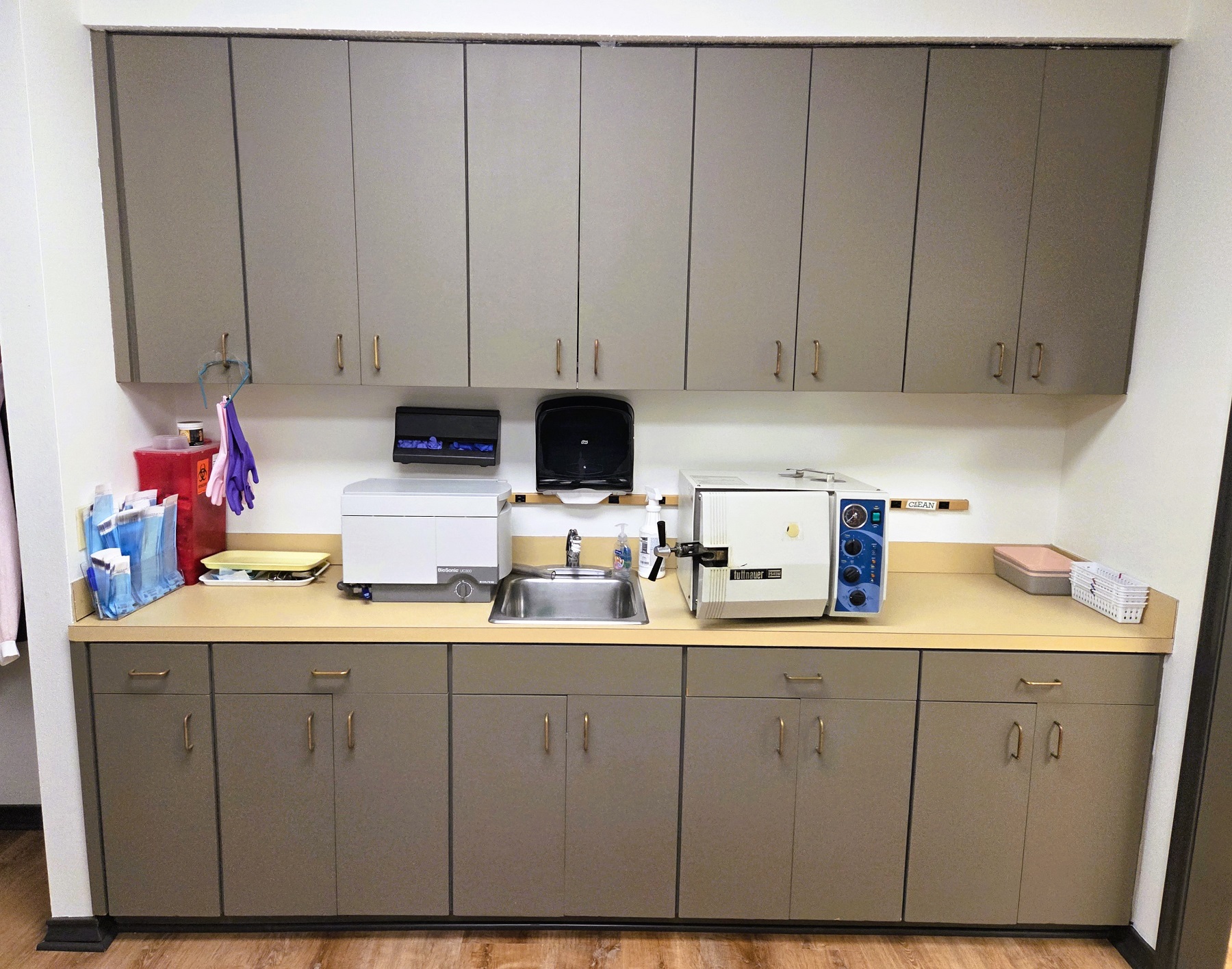 light walls with dark stained wood baseboards. Dark cabinetry with orangy counter tops and a stainless steal sink, hold dental sterilization equipment