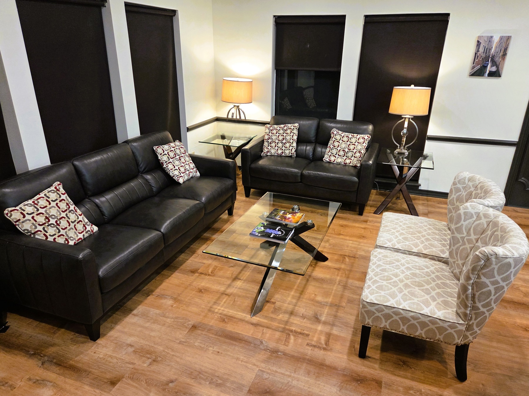 Light walls with dark stained wood baseboards and medium stained wood flooring. A set of black cushioned sofa, loveseat, and a pair of light patterned chairs surround a glass coffee table.