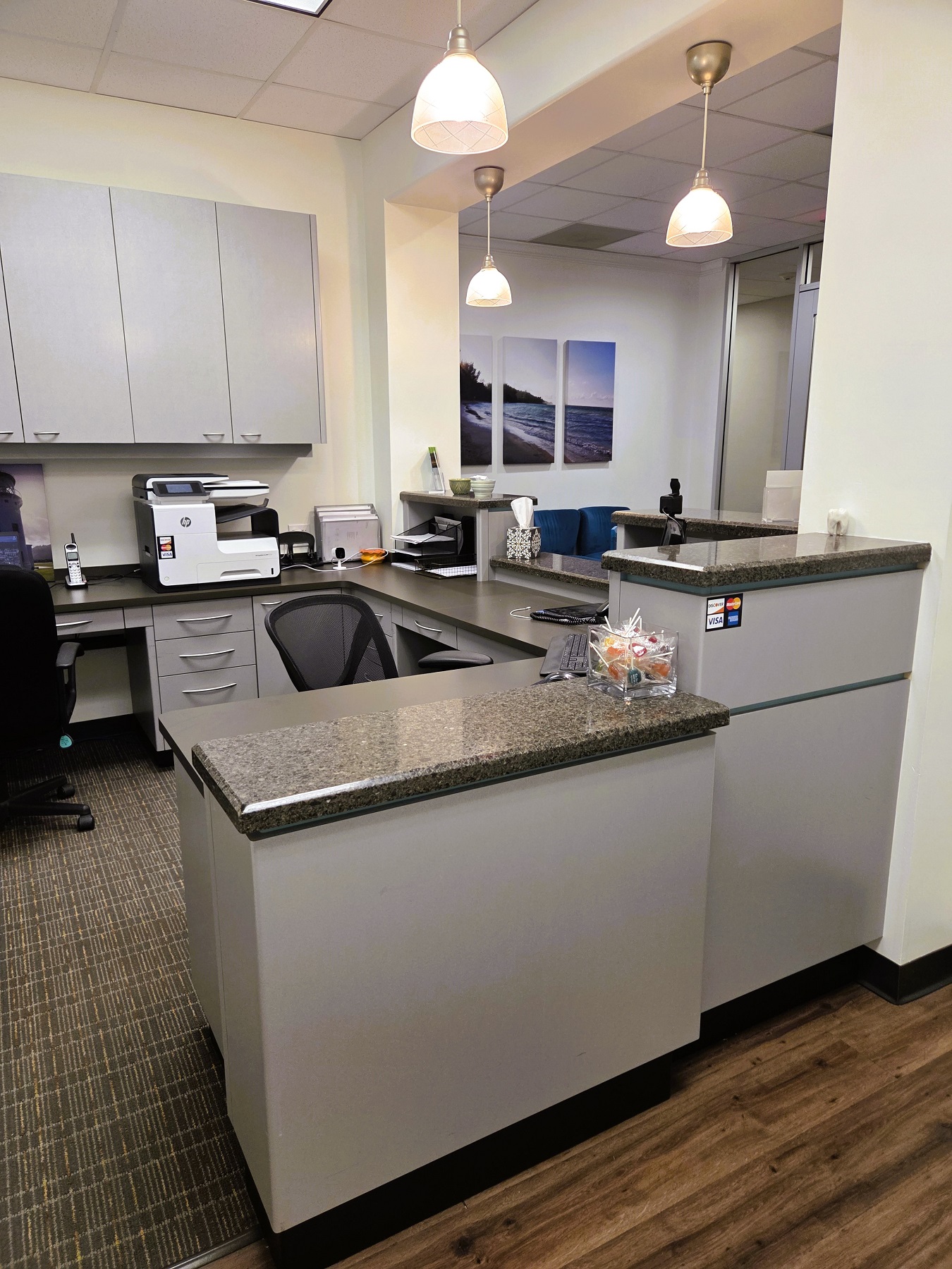 white walls with black baseboards and dark wood flooring. Commercial carpet is in the front office area. Grey builtin cabinetry with darker countertops hold lots of black office equipment, tools, and supplies.
