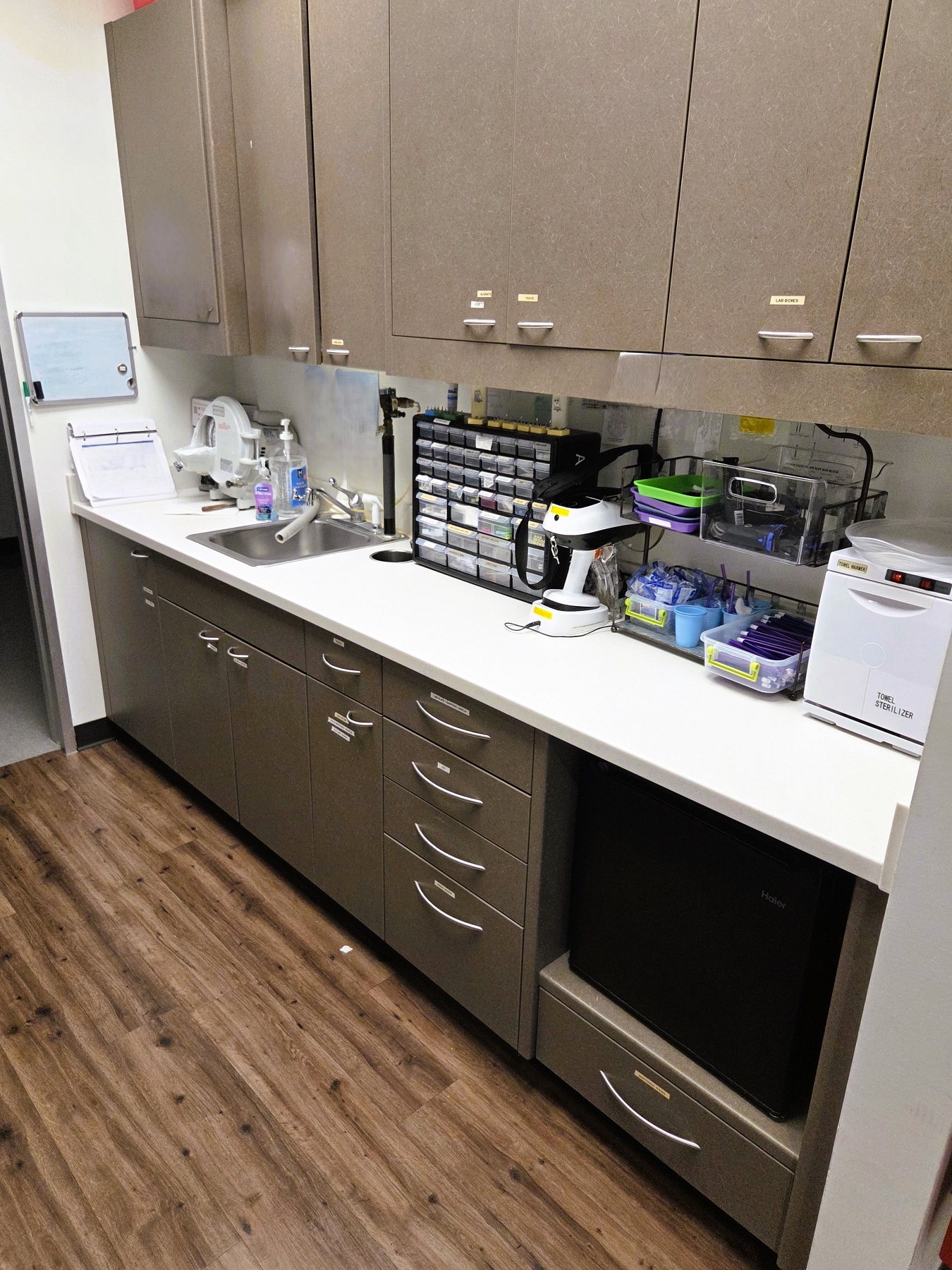 White walls with black baseboards and stained wood flooring. Dark cabinetry with white counter tops hold lots of dental equipment, tools, and supplies.