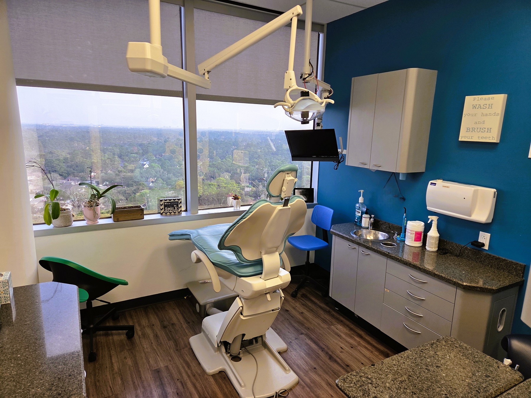 White walls with black baseboards and medium stained wood flooring. Large picture windows with a beautiful view. Dental procedural chair anchors the room with overhead dental lighting. Builtin grey cabinetry with darker countertops hold lots of dental equipment, tools, and supplies.