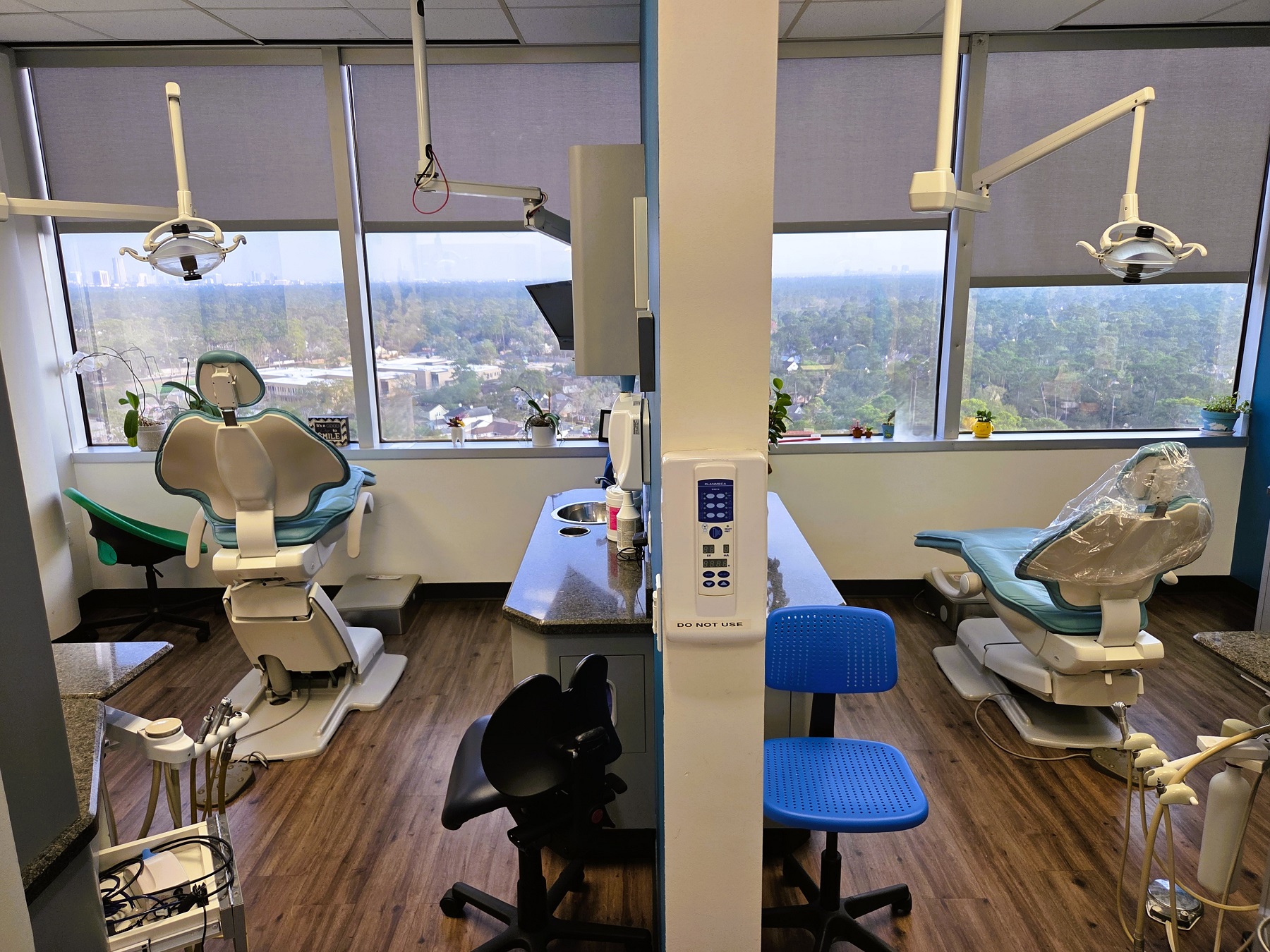 White walls with black baseboards and medium stained wood flooring. Large picture windows with a beautiful view. Dental procedural chair anchors the room with overhead dental lighting. Builtin grey cabinetry with darker countertops hold lots of dental equipment, tools, and supplies.