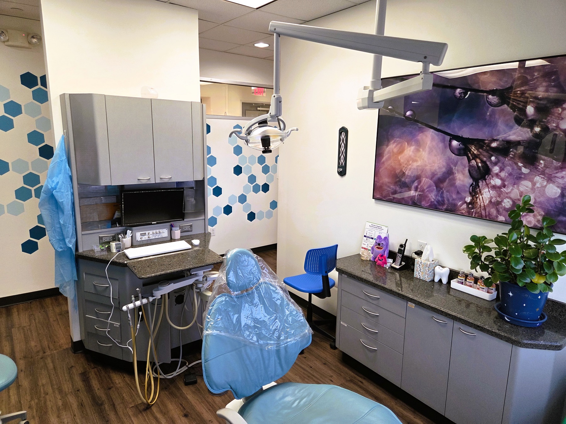 White walls with black baseboards and medium stained wood flooring. Dental procedural chair anchors the room with overhead dental lighting. Builtin grey cabinetry with darker countertops hold lots of dental equipment, tools, and supplies.