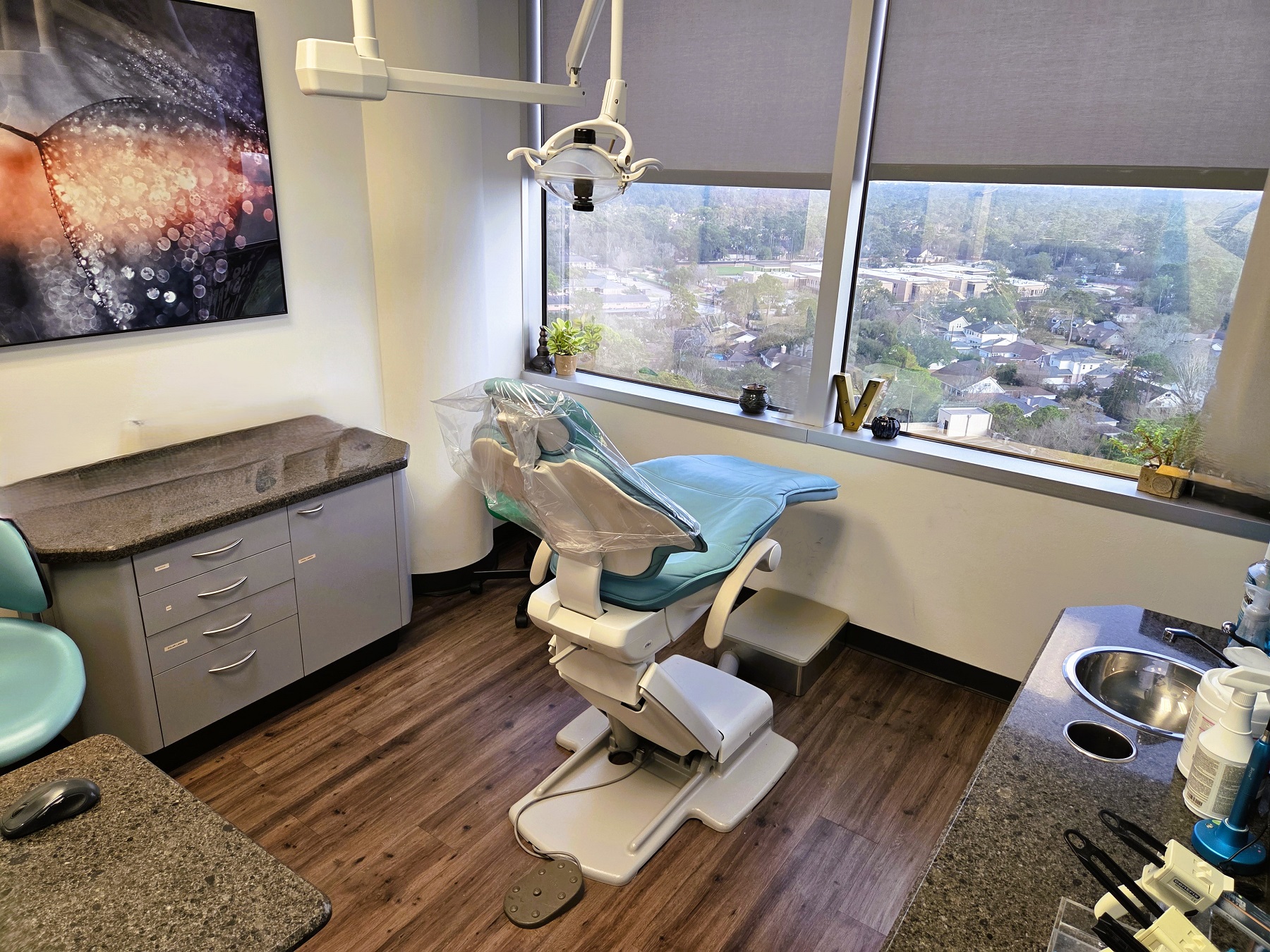 White walls with black baseboards and medium stained wood flooring. Large picture windows with a beautiful view. Dental procedural chair anchors the room with overhead dental lighting. Builtin grey cabinetry with darker countertops hold lots of dental equipment, tools, and supplies.