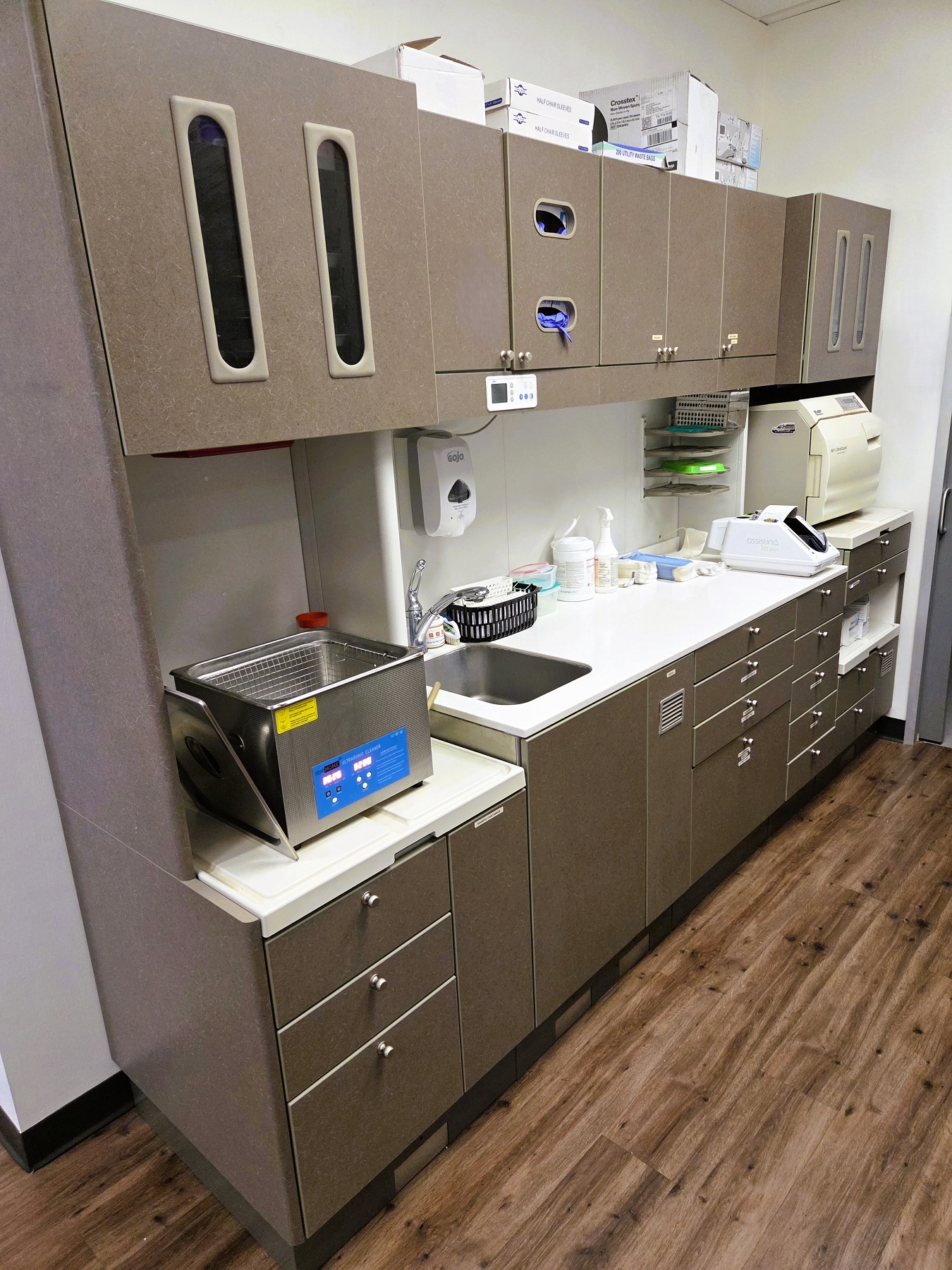 White walls with black baseboards and stained wood flooring. Dark cabinetry with white counter tops hold lots of dental equipment, tools, and supplies.