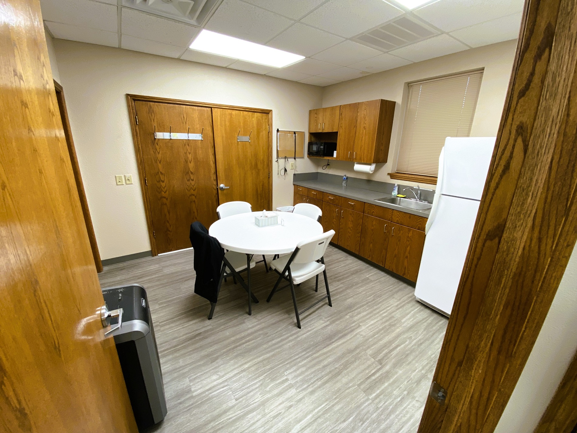 Tan walls with dark baseboards and grey wood-look flooring. Dark stained cabinetry with dark grey countertop and a stainless steel sink sit next to a white refrigerator. Around white plastic table is surrounded by white plastic chairs