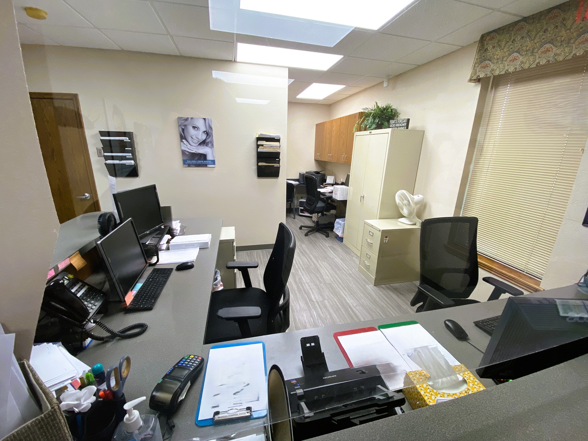 Tan walls with dark baseboards. Grey wood-look flooring. Builtin "L" shaped desk with a grey desktop holds lots of office equipment and supplies. A couple of black desk chairs sit at the desk. There's a couple of large tan file cabinets sitting against the all in front of a small desk in the background. It has additional office equipment and an office chair.