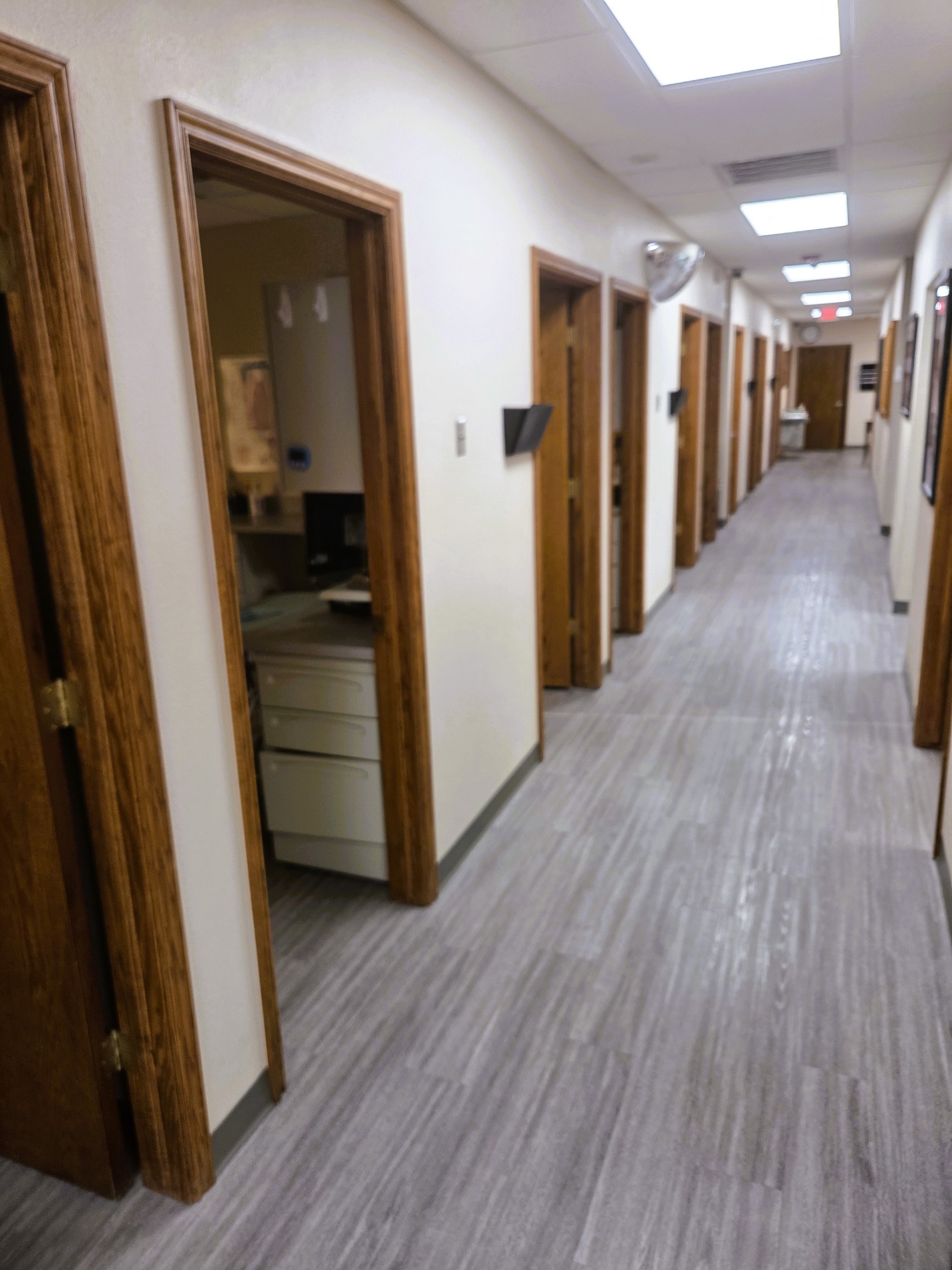 Tan walls with black baseboards and grey, wood-grain, wood-look flooring. Stained wood door jams line the hallway and lead to the various operatories and rooms of the practice.