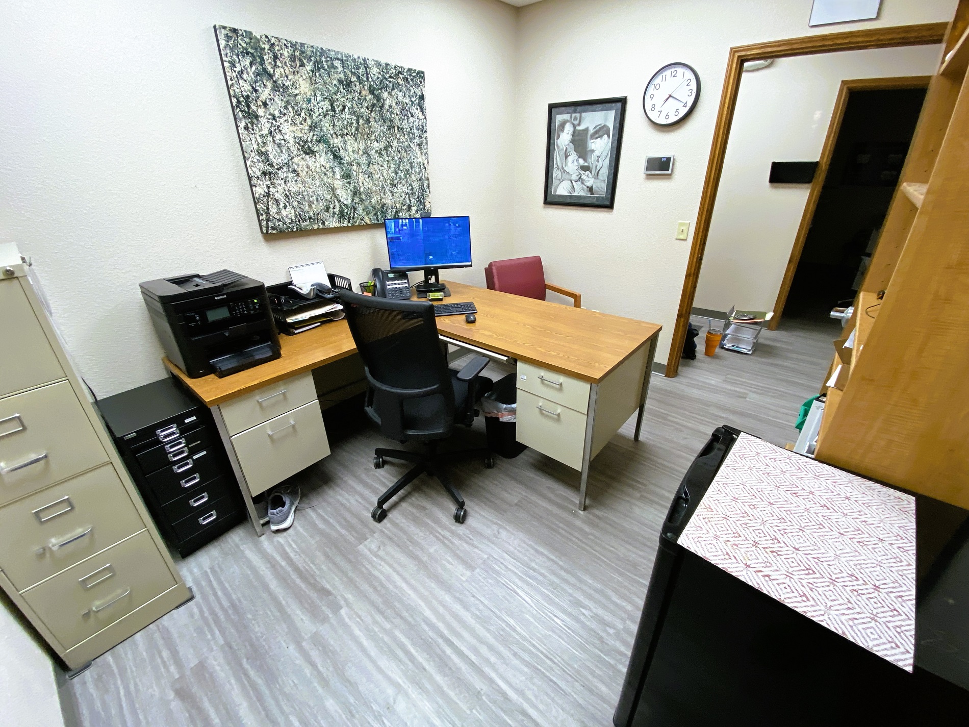 Tan walls with dark baseboards and grey wood-look flooring. Random furniture fills the room. There's a couple file cabinets, a wood bookshelf, a large framed printo on the wall, and a small "L" shaped desk with black rolling office chair. Various office equipment and supplies sit on the desk.