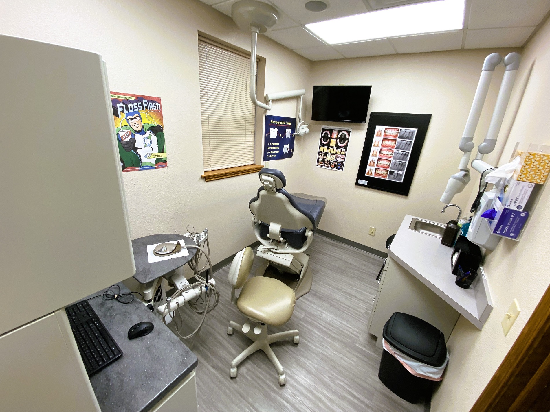 Tan walls with dark baseboards. Posters on the wall. Grey wood-look flooring. Dental procedural chair anchors the room with overhead dental procedural light. A small dark desk with overhead cabinet holds office equipment and dental supplies and tools.