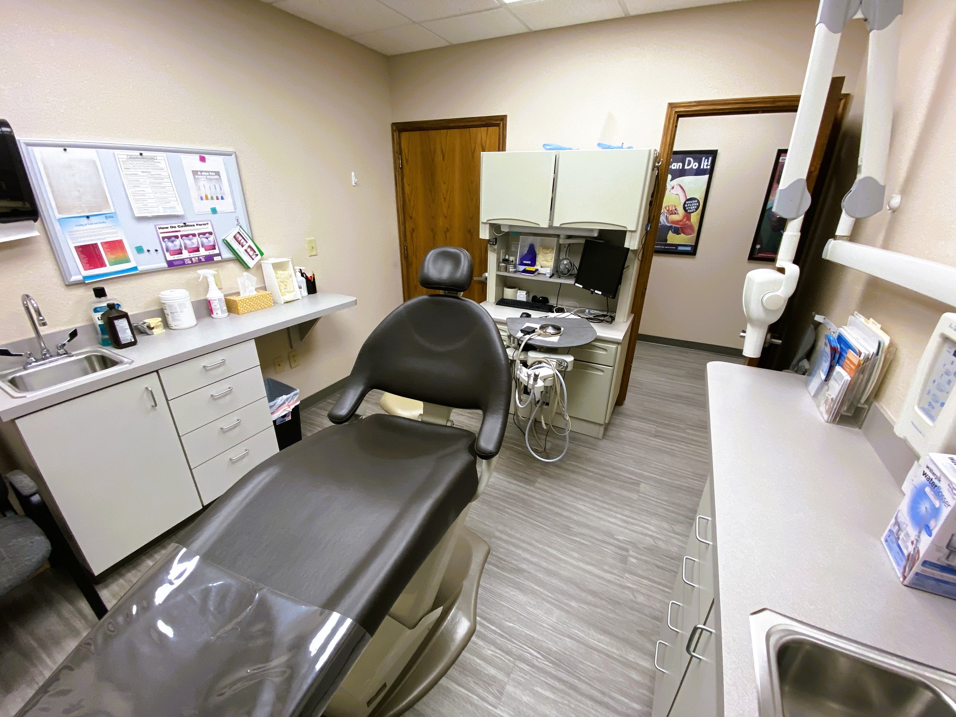 Tan walls with dark baseboards. Posters on the wall. Grey wood-look flooring. Dental procedural chair anchors the room with overhead dental procedural light. A small dark desk with overhead cabinet holds office equipment and dental supplies and tools.