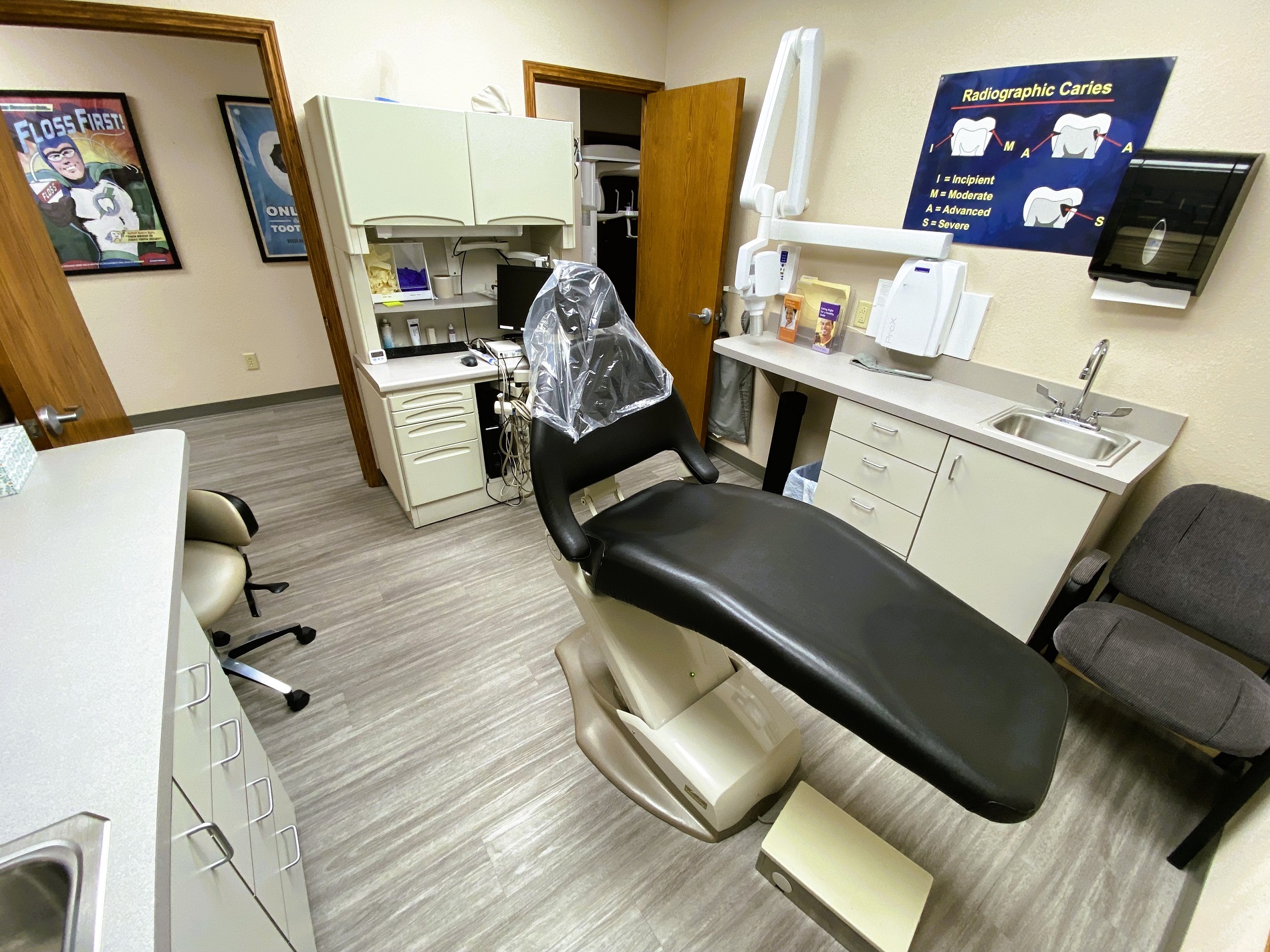 Tan walls with dark baseboards. Posters on the wall. Grey wood-look flooring. Dental procedural chair anchors the room with overhead dental procedural light. A small dark desk with overhead cabinet holds office equipment and dental supplies and tools.