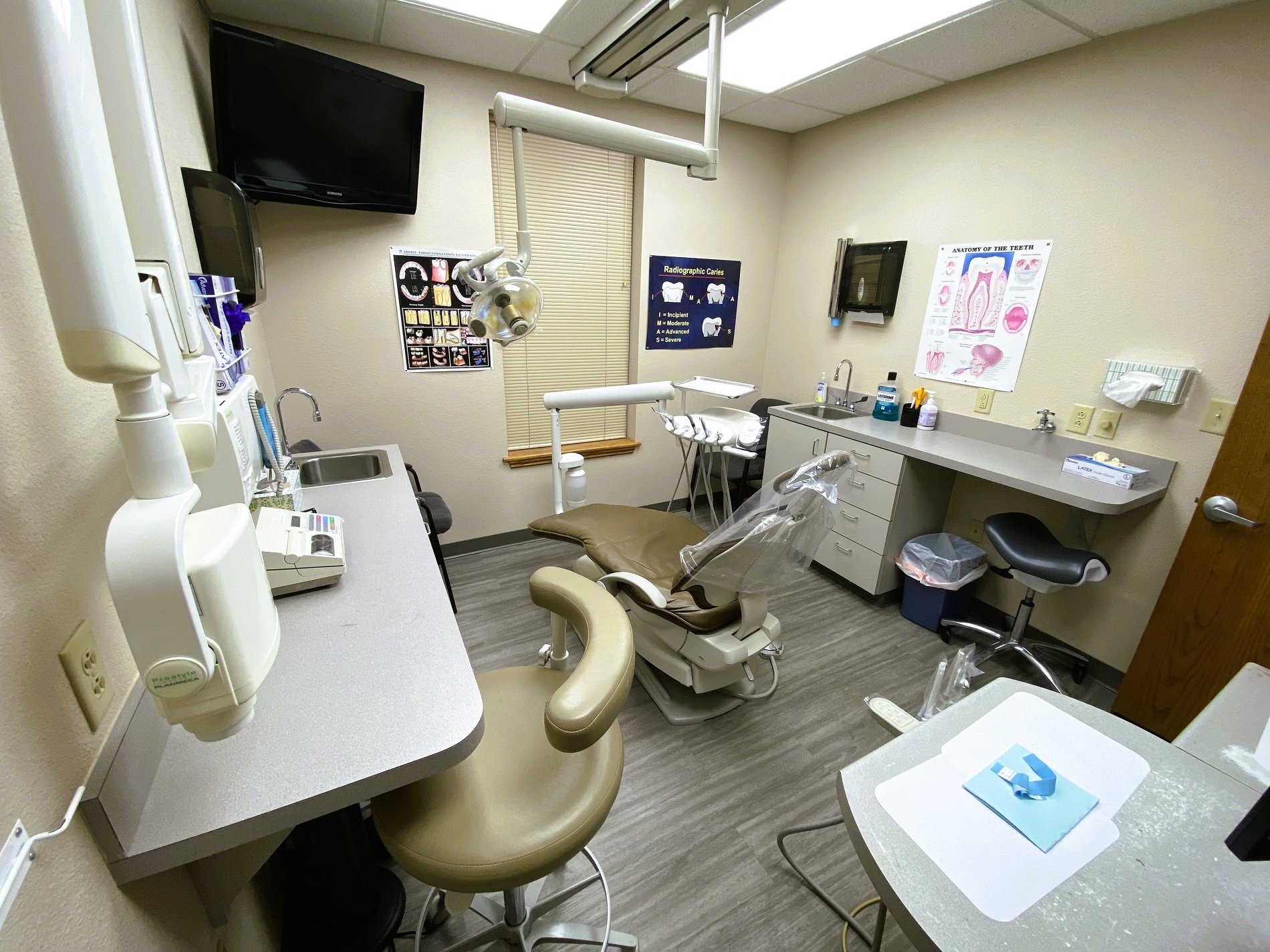 Tan walls with dark baseboards. Posters on the wall. Grey wood-look flooring. Dental procedural chair anchors the room with overhead dental procedural light. A small dark desk with overhead cabinet holds office equipment and dental supplies and tools.
