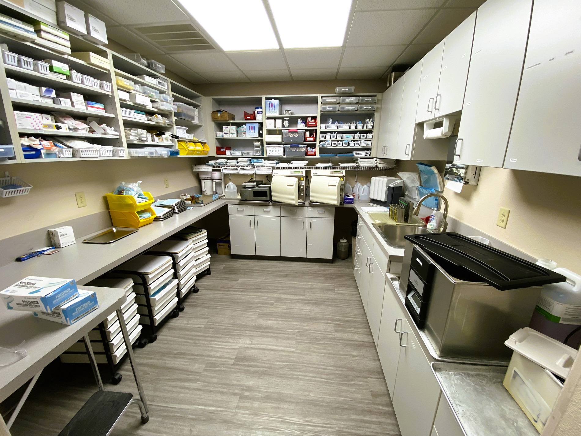 Tan walls with dark baseboards. Grey wood-look flooring. Huge grey built-in desk and shelves with a light grey countertop and stainless steel sink. Dental tools, equipment, and supplies fill the counter top, shelves, and cabinets.