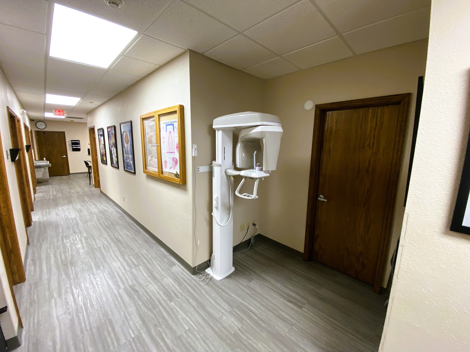 Tan walls with dark baseboards. Grey wood-look flooring. Framed posters and artwork lines the walls down the hallway. Large dental x-ray machine sits in an alcove off the hallway.