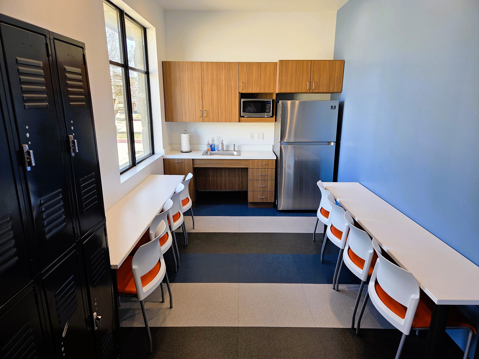 Light walls with one blue accent wall, dark baseboards, and two tone white and blue tile flooring. A large window lets in lots of natural light. Black lockers along part of one wall allow security for personal belongings. A counter top and 4 cushioned chairs line tow of the walls.