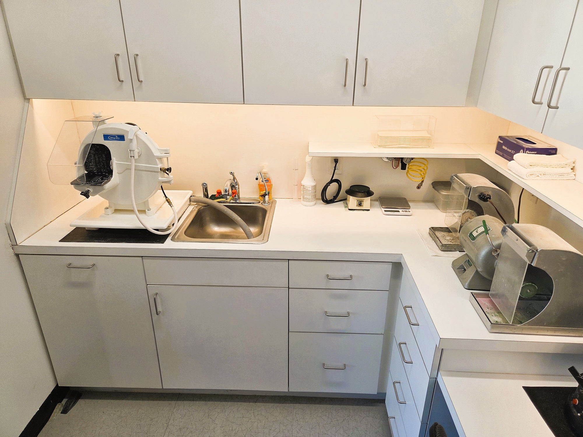 Light walls with dark baseboards and light tile flooring. White cabinets, both uppers and lowers, with white countertops with a builtin single basin stainless steel sink hold lots of dental lab equipment supplies, tools, and equipment.