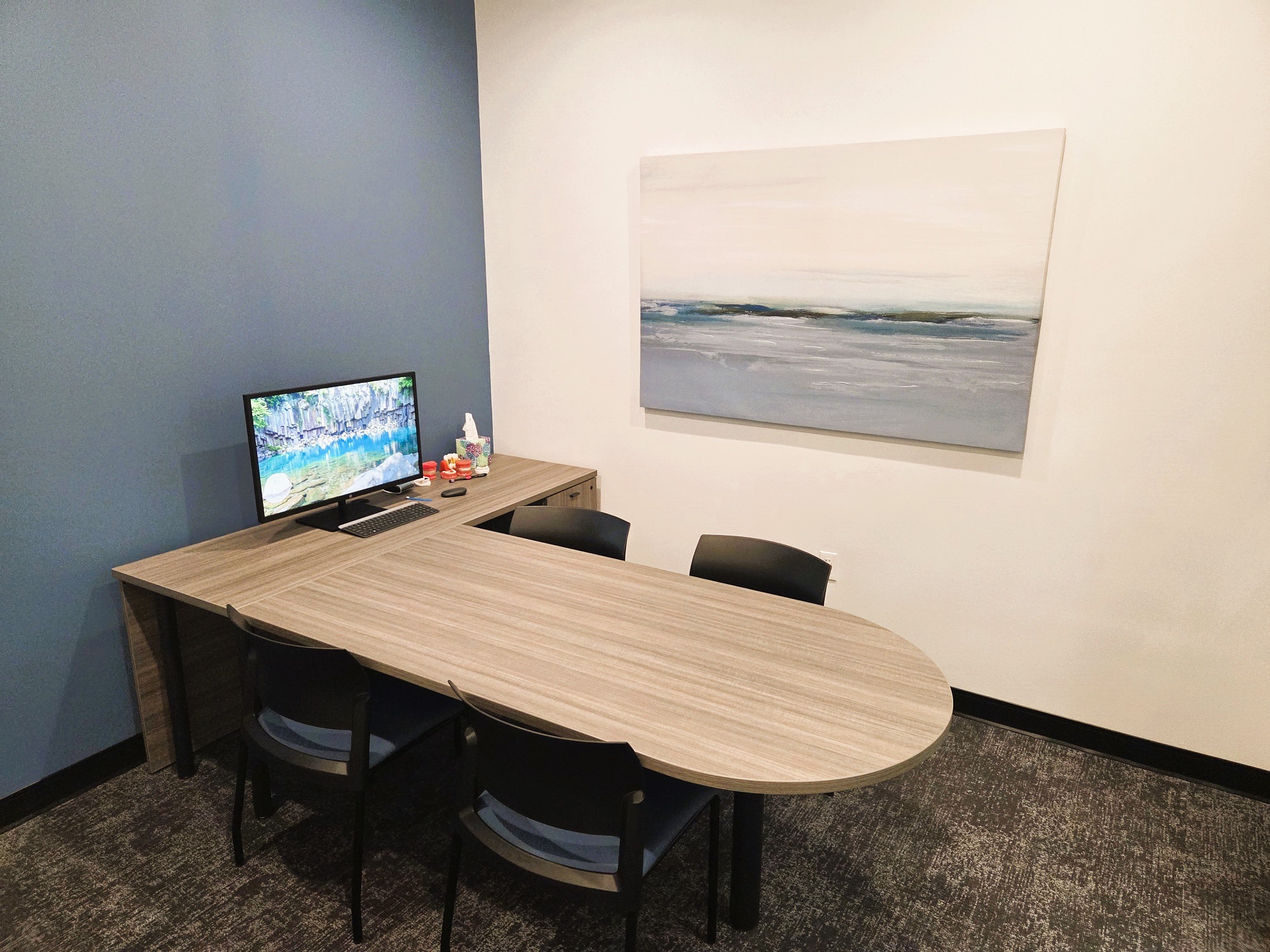 One light grey and one dark grey wall creates contrast and interest in this consultation office. Short pile dark grey carpet is warm and inviting with a warm L-shaped wood desk having several chairs around it holding a computer monitor.