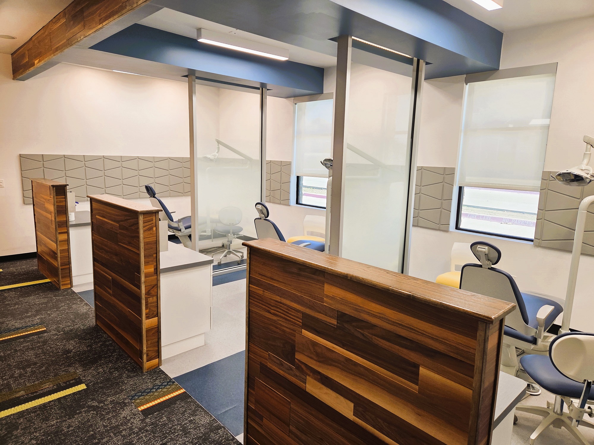 Light grey walls with darker grey feature tiles running horizontally around the room. Glass partitions allow the natural light from large windows to go into all of the dental operatories. Two tone epoxy flooring make cleanup in the dental operatories super easy while darker grey commercial carpet makes the walkways feel warm and welcoming. The horizontal wood features between the dental operatories carries throughout the practice. Dark blueish-grey dental procedural chairs anchor each operatory with overhead dental procedural lighting. A small cabinet on the inside of the wood partition holds lots of dental equipment and supplies. A rolling chair is in each of the operatories for the dentist or dental assistant.