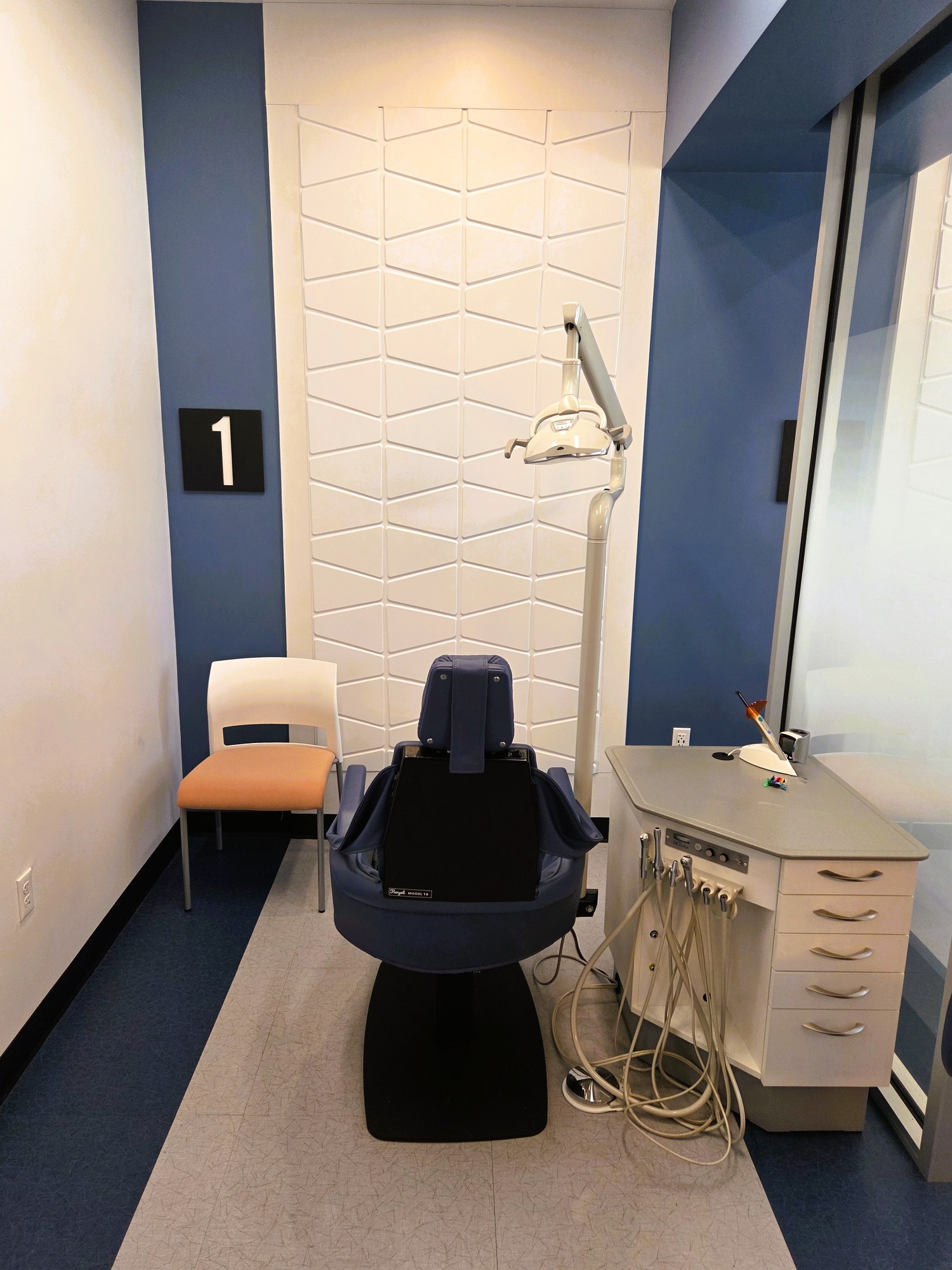 Two tone white and blue walls including a white tile feature wall in the middle of the Ortho bay. Black baseboards and floor tiles that match the two tone walls. A dental procedural chair anchors the ortho bay. Along with overhead dental lighting and a white cabinet that houses all of the needed dental equipment and supplies.