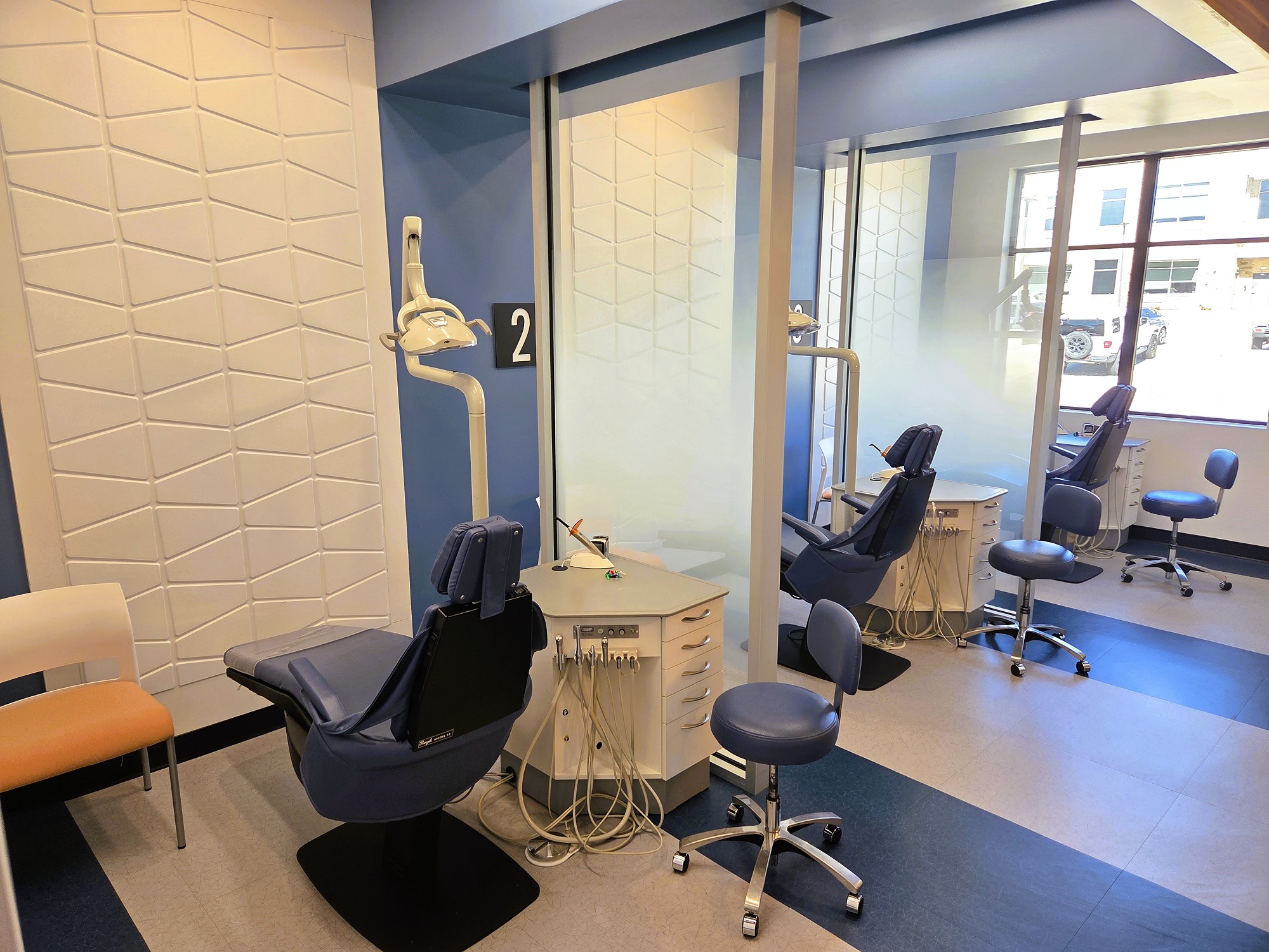 Two tone white and blue walls including a white tile feature wall in the middle of the Ortho bay. Black baseboards and floor tiles that match the two tone walls. A dental procedural chair anchors the ortho bay. Along with overhead dental lighting and a white cabinet that houses all of the needed dental equipment and supplies.