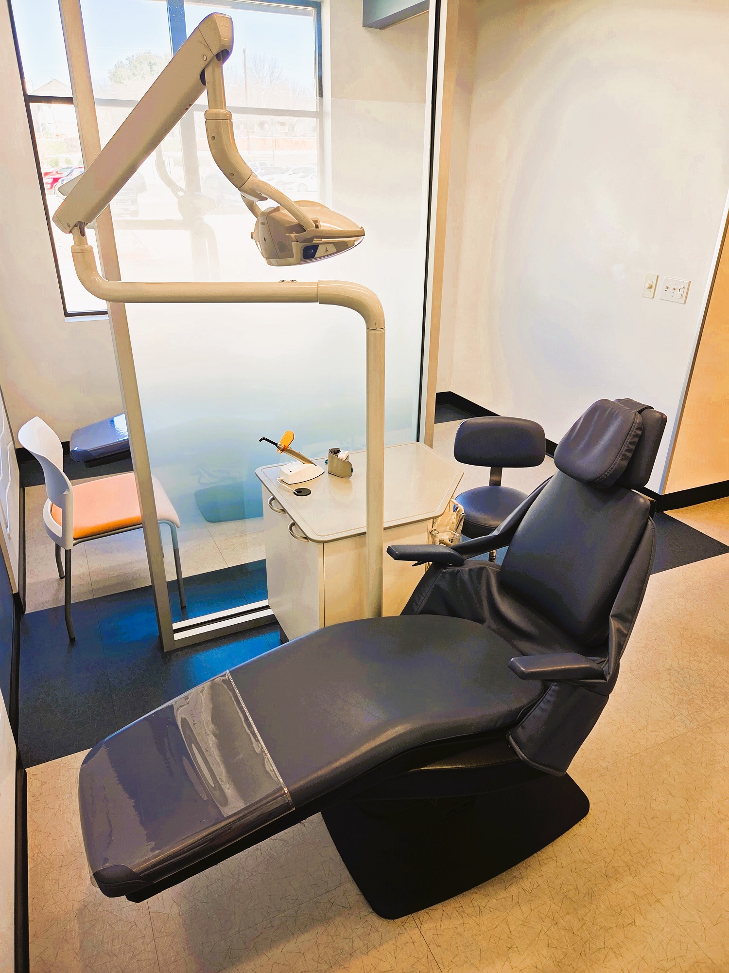 Two tone white and blue walls including a white tile feature wall in the middle of the Ortho bay. Black baseboards and floor tiles that match the two tone walls. A dental procedural chair anchors the ortho bay. Along with overhead dental lighting and a white cabinet that houses all of the needed dental equipment and supplies.