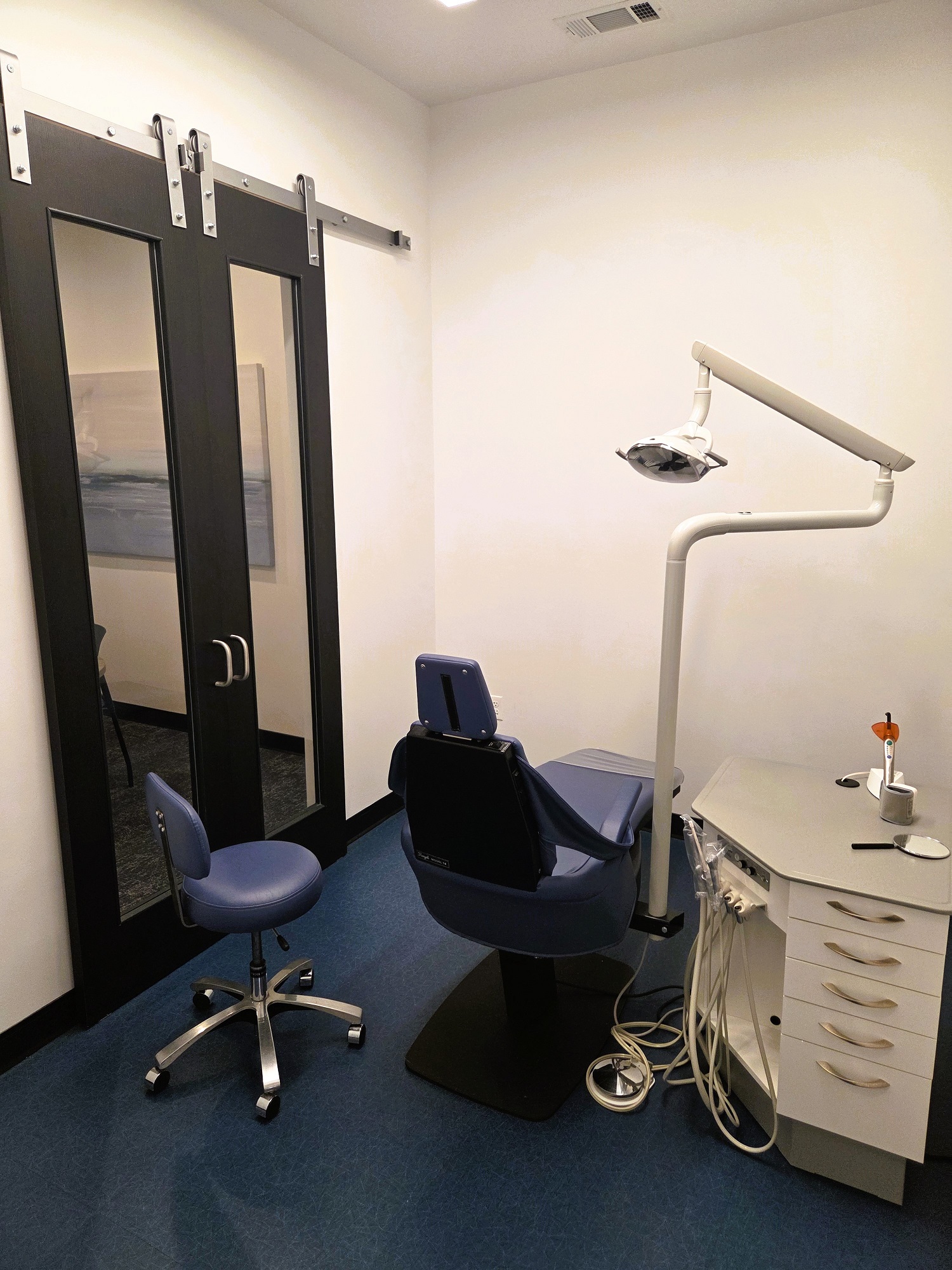 Light walls with dark trim, doors, and baseboards. Dark blue tile flooring make rolling chairs easy to move. A dark dental procedural chair anchors the room, with an overhead dental light and a small cabinet that holds lots of dental tools and equipment.