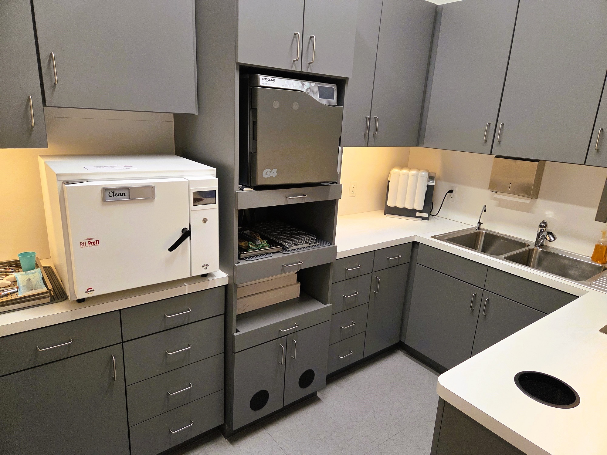 light walls with dark baseboards and light tile flooring. Dark grey built-in cabinetry with light countertops holds lots of dental sterilization equipment, tools, and supplies.