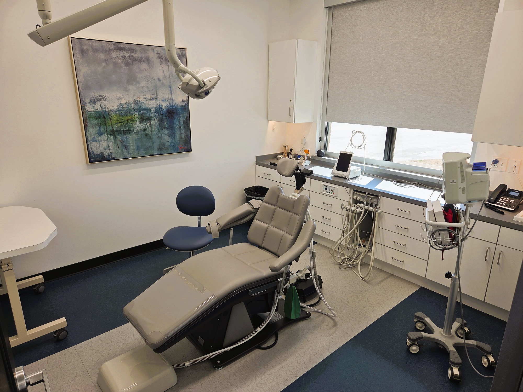Light walls with large framed artwork, dark baseboards and two tone tile flooring. Light grey dental procedural chair anchors the room with a rolling chair for the surgeon. A large built-in white cabinet houses all of the needed dental equipment, tools, and supplies.