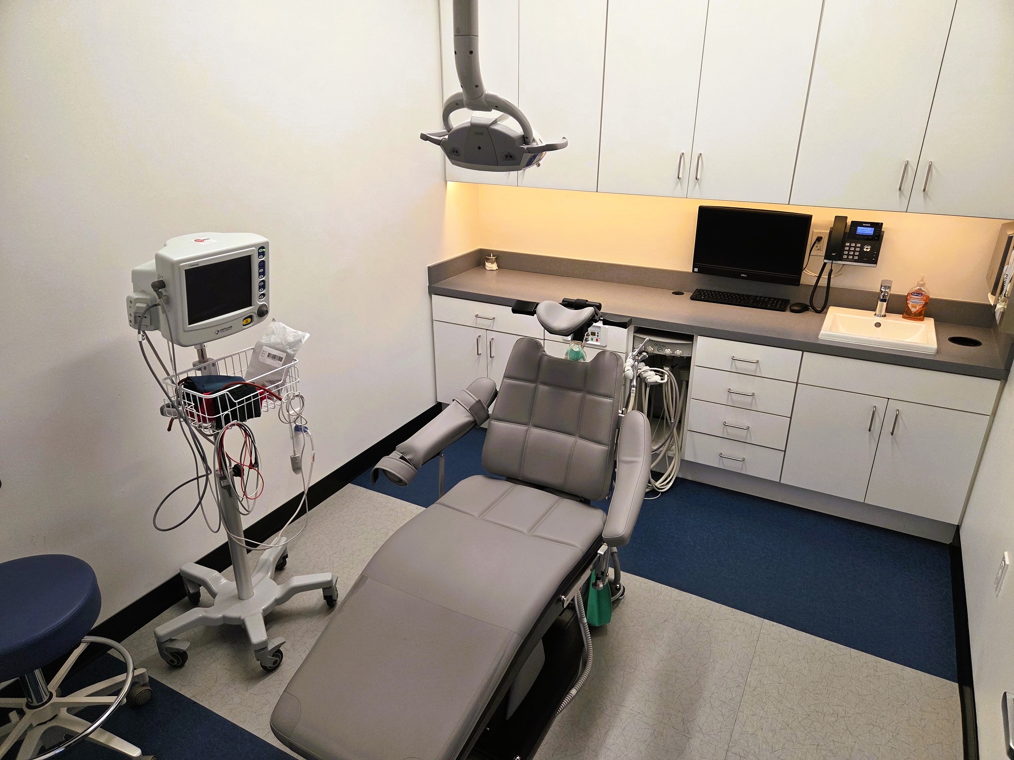 Light walls with large framed artwork, dark baseboards and two tone tile flooring. Light grey dental procedural chair anchors the room with a rolling chair for the surgeon. A large built-in white cabinet houses all of the needed dental equipment, tools, and supplies.