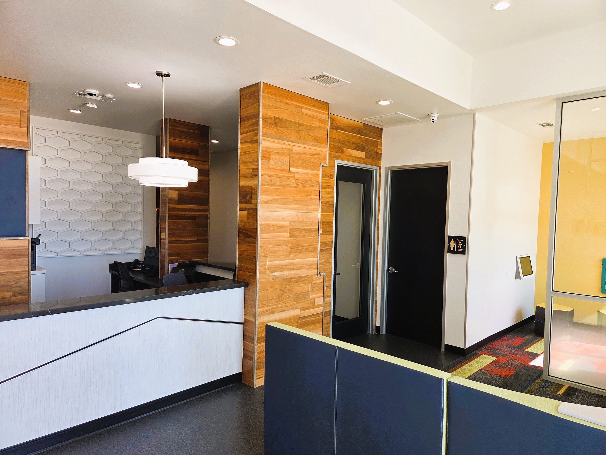 Light grey walls with black baseboards and epoxy flooring. Grey tile feature wall in the very back behind the reception desk and horizontal stained wood plank on several of the columns and walls around the reception desk. A dark counter top on the builtin reception desk with dark office furniture behind. A very light and bright waiting area with cushioned seats sits next to floor to ceiling windows.