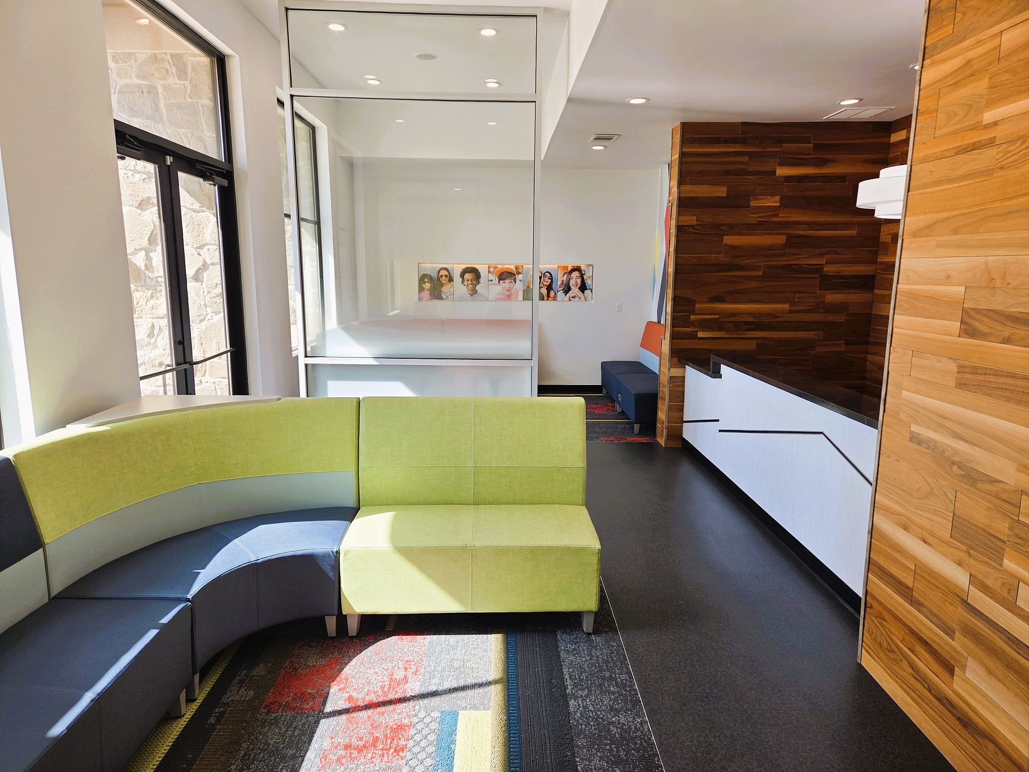 Light grey walls with black baseboards and epoxy flooring. Horizontal stained wood plank on several of the columns and walls around the reception desk. A dark counter top on the builtin reception desk with dark office furniture behind. A very light and bright waiting area with cushioned seats sits next to floor to ceiling windows.