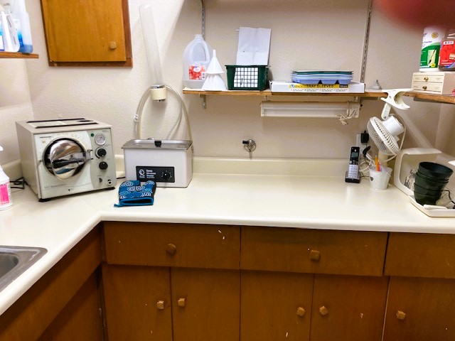 Tan walls with medium stained cabinetry and adjustable shelving, with white counter tops. Lots of dental lab equipment, tools, and supplies and a stainless steel sink.