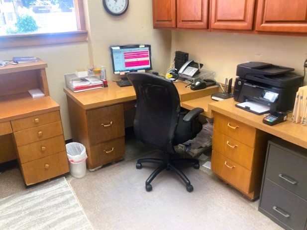 Tan walls with black base boards and a medium stained wood trimmed window allowing lots of natural light into the room. Matching medium stained wood desks line the walls with matching wood upper cabinets on one wall. There is a black rolling office chair and lots of office equipment on the desks.
