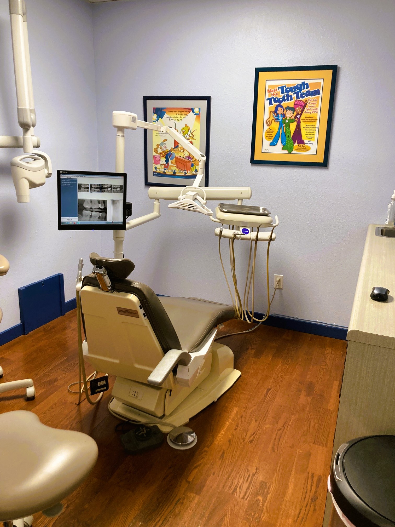 Light blue walls with black base boards and hardwood flooring. Tan dental procedural chair anchors the room with matching rolling chair. Overhead dental procedural lighting and an x-ray machine mounted on the wall. Framed posters are on the other wall and a tan cabinet holds tons of dental supplies, tools, and equipment.