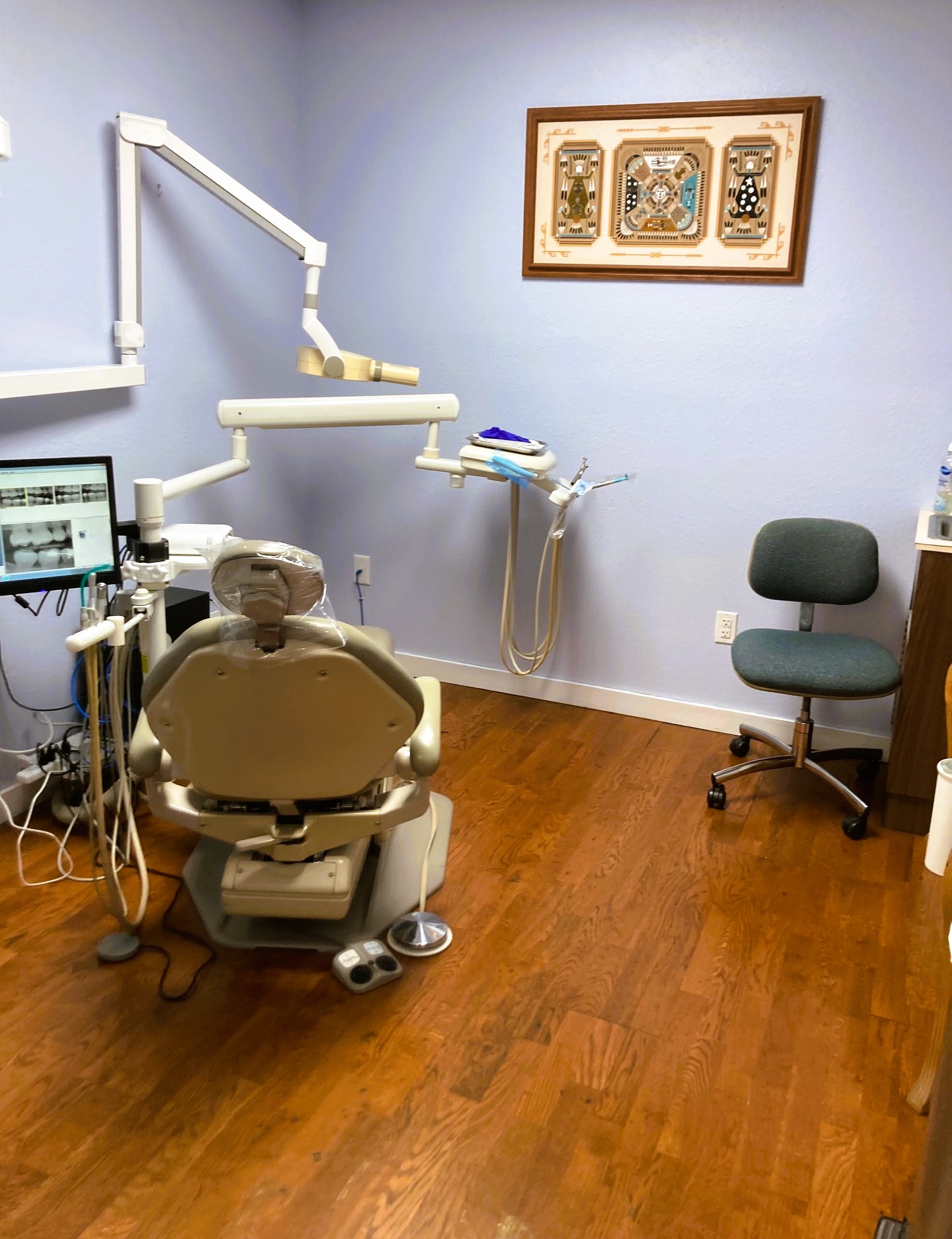 Light blue walls with black base boards and hardwood flooring. Tan dental procedural chair anchors the room with matching rolling chair. Overhead dental procedural lighting and an x-ray machine mounted on the wall. Framed posters are on the other wall and a tan cabinet holds tons of dental supplies, tools, and equipment.