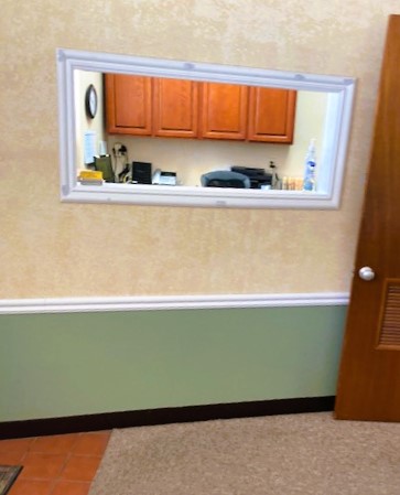 Two tone wall, tan on top and medium green below the white chair rail with a black base board at the bottom. Short, tan commercial carpet on the floor. White framing around the reception window looking back into the front office. A wood door leads to the back of the practice.