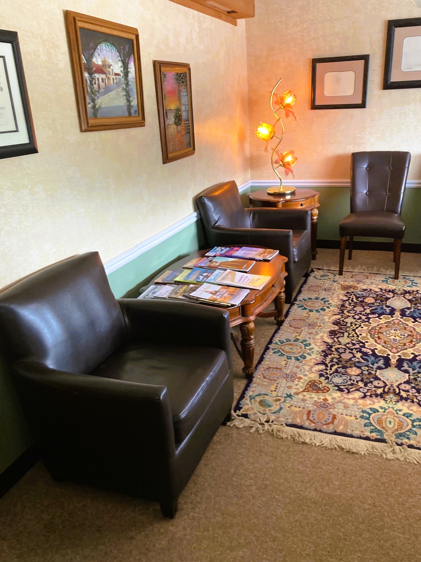 Two tone walls, light upper and medium green lower below the white chair rail and black base board. Short, tan commercial carpet is covered with an ornamental rug. Black, leather, cushioned chairs line the room backing up to the wall and to a huge window on the other. The window allows lots of natural light to flow into the room. A ceiling fan with a light are centered in the room above the rug. There is a small side table between some of the chairs and in front of the window that holds a globe. Framed certificates and diplomas line the wall.
