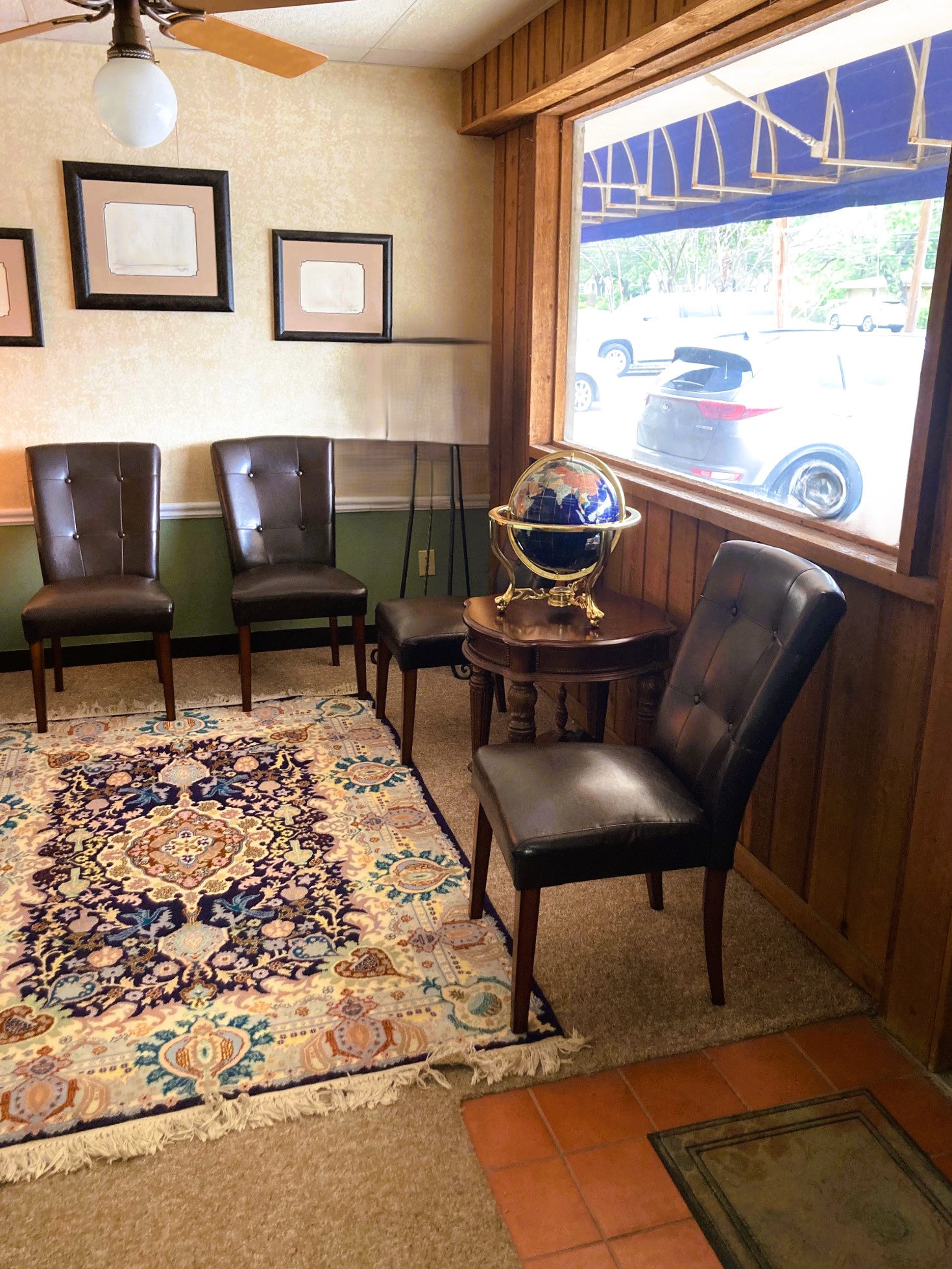Two tone walls, light upper and medium green lower below the white chair rail and black base board. Short, tan commercial carpet is covered with an ornamental rug. Black, leather, cushioned chairs line the room backing up to the wall and to a huge window on the other. The window allows lots of natural light to flow into the room. A ceiling fan with a light are centered in the room above the rug. There is a small side table between some of the chairs and in front of the window that holds a globe. Framed certificates and diplomas line the wall.