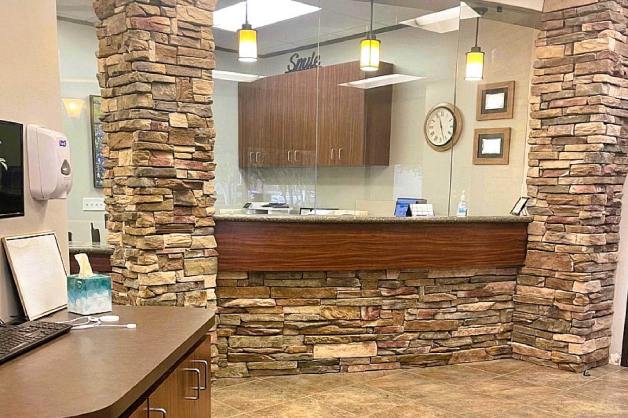 White walls with dry stacked stone accents and large brown tile flooring. Natural stained wood built in desk with huge glass windows looking into the front office with curved desk tops. Framed diplomas and certificates on the wall around a natural wood stained upper cabinet to hold office supplies and equipment. A round clock on the wall and several pendant lights hang over the front desk.