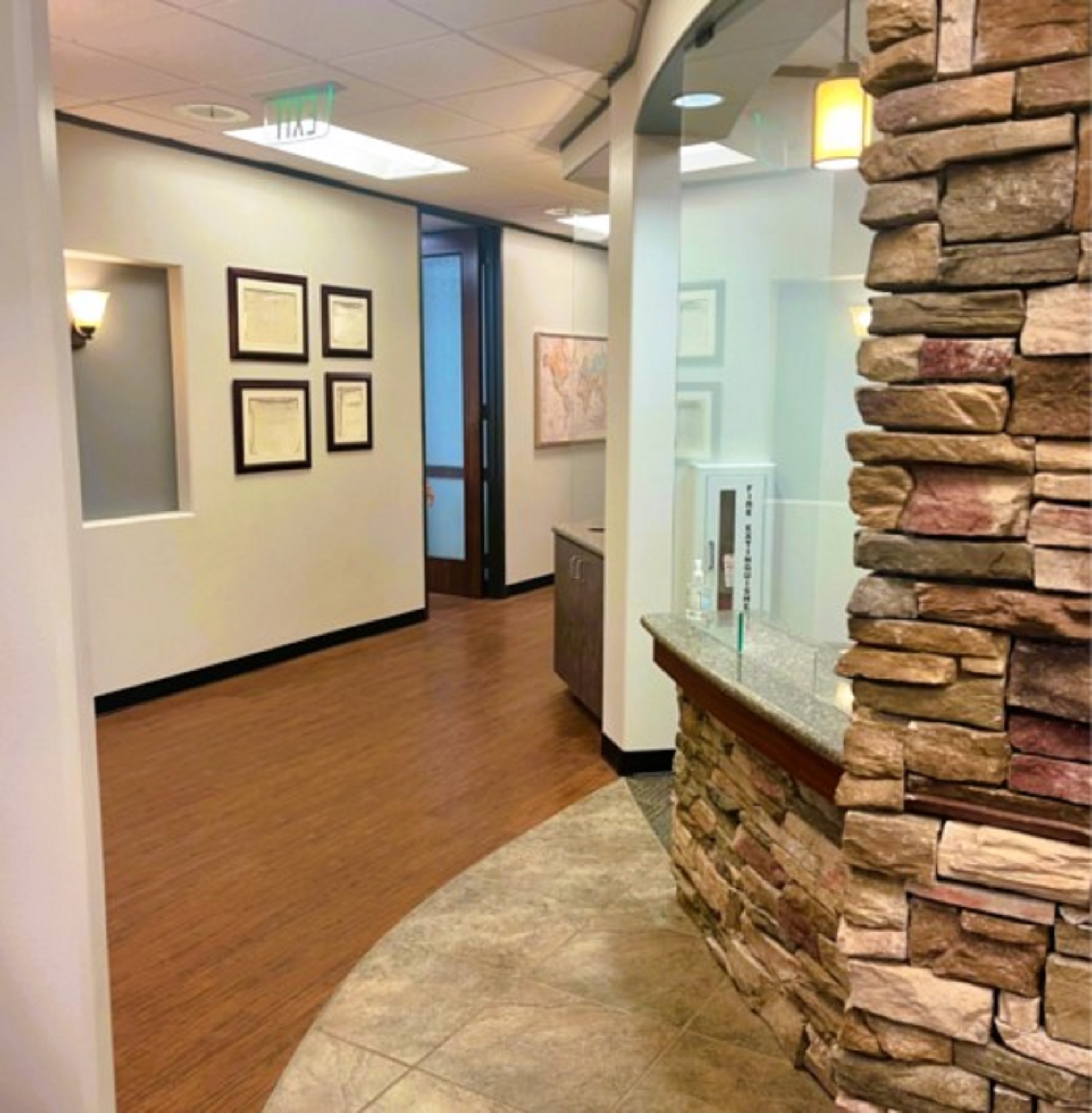 light walls with black baseboards, and framed diplomas and artwork. Dry stacked stone accent walls and wall sconces bring character to the hallway. Large brown tile flooring just in a curve to blend into the wood-look vinyl flooring that runs the length of the hallways. Large framed doorways with stained wood framed doors with opaque glass for privacy and light.