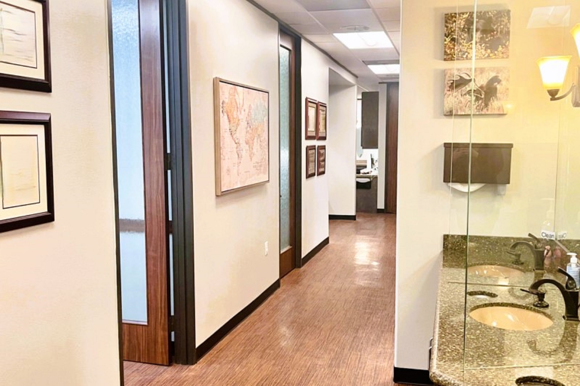 light walls with black baseboards, and framed diplomas and artwork. Dry stacked stone accent walls and wall sconces bring character to the hallway. Large brown tile flooring just in a curve to blend into the wood-look vinyl flooring that runs the length of the hallways. Large framed doorways with stained wood framed doors with opaque glass for privacy and light.
