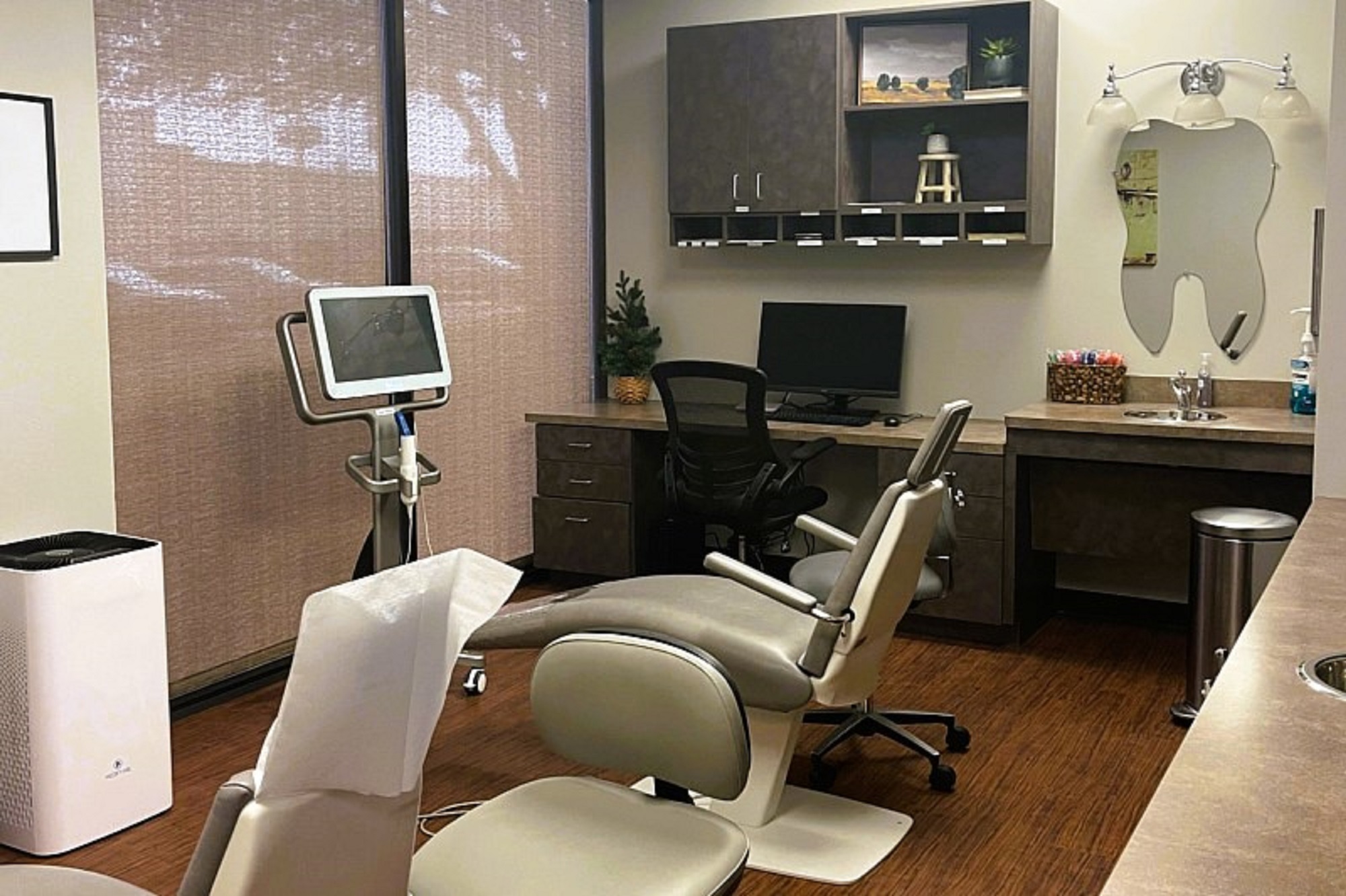 Light walls with dark baseboards and dark brown commercial carpet tile flooring. A large floor to ceiling window is covered by a cloth blind. Dental procedural chairs with matching rolling chairs and equipment stands create a wonderful dental hygiene operatory.