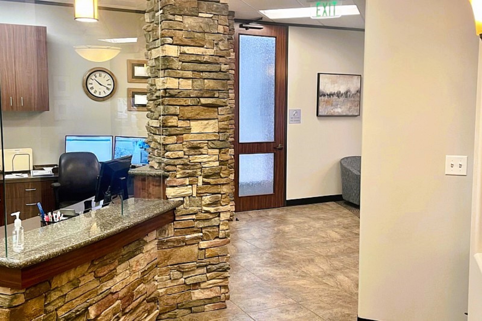 White walls with dry stacked stone accents and large brown tile flooring. Natural stained wood built in desk with huge glass windows looking into the front office with curved desk tops. Framed diplomas and certificates on the wall around a natural wood stained upper cabinet to hold office supplies and equipment. A round clock on the wall and several pendant lights hang over the front desk.