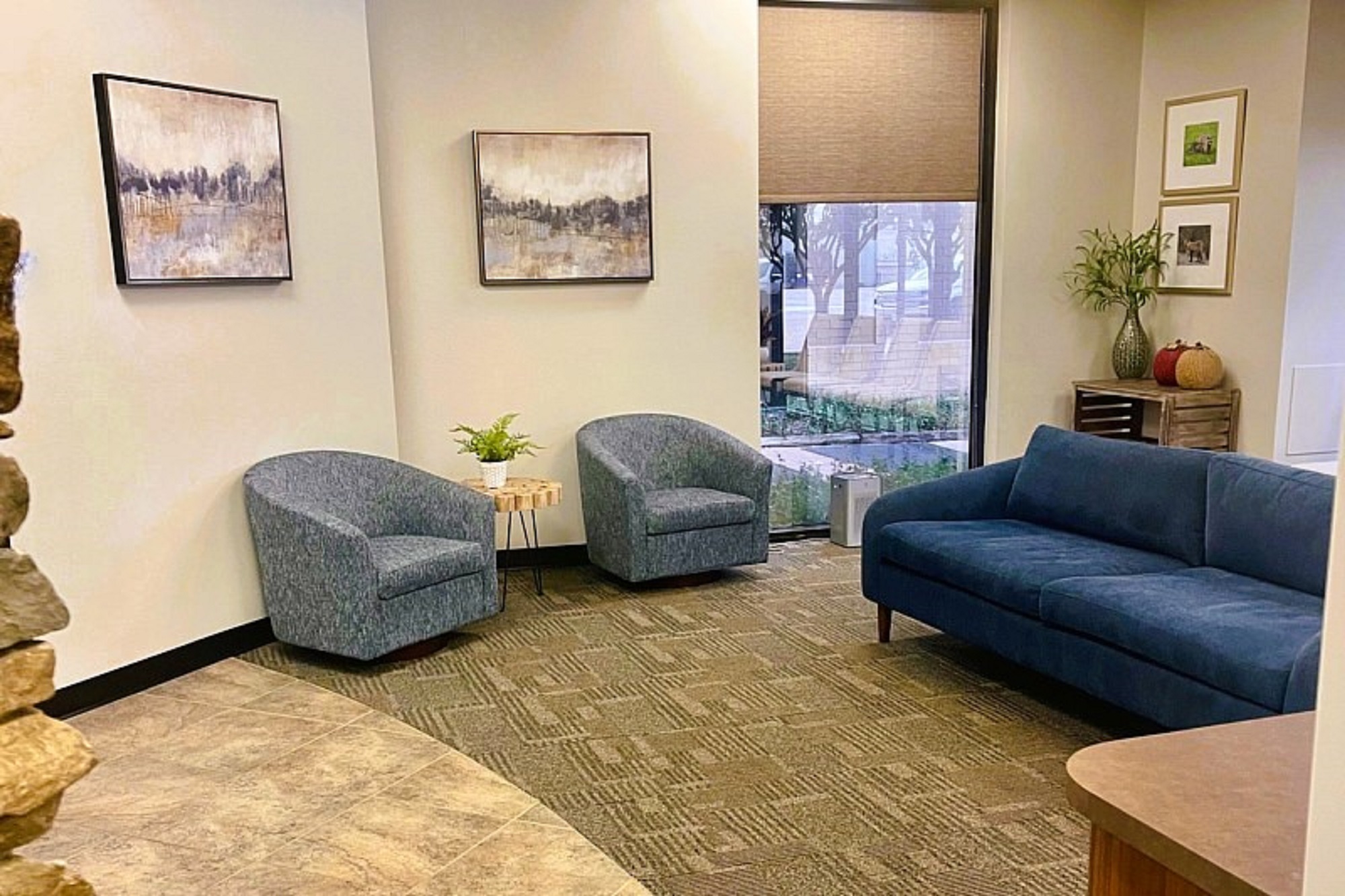 White walls and a tan accent wall with black base boards. Framed artwork on the walls on either side of a floor to ceiling window with a fabric Roman shade. Rock accent walls with large brown floor tiles on the left and textured commercial carpet tiles in the waiting area. Light blue cushioned chairs and a dark blue cushioned sofa in MCM style provide a comfortable place for patients to wait.