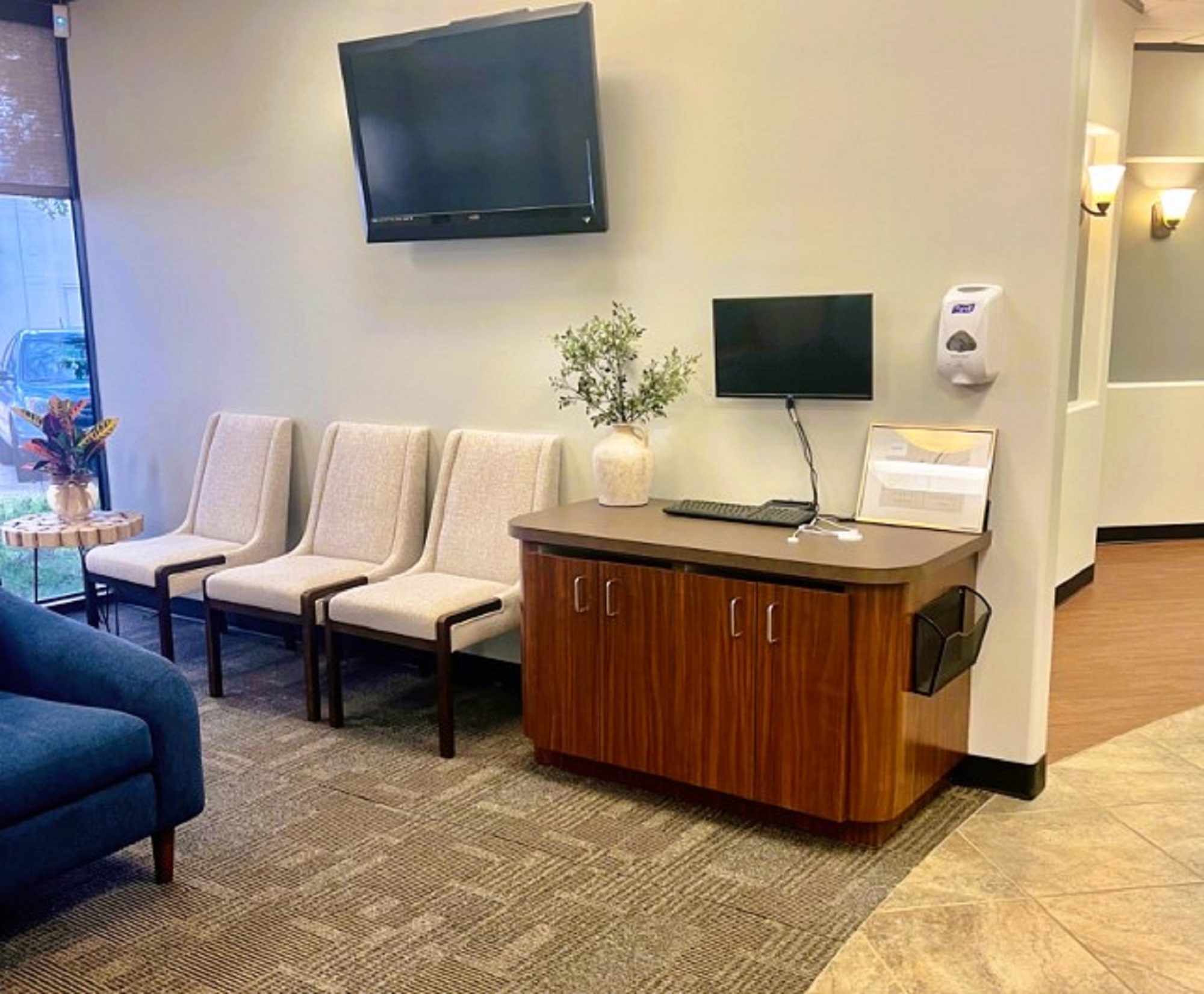 White walls and a tan accent wall with black base boards. Framed artwork on the walls on either side of a floor to ceiling window with a fabric Roman shade. Rock accent walls with large brown floor tiles on the left and textured commercial carpet tiles in the waiting area. Light blue cushioned chairs and a dark blue cushioned sofa in MCM style provide a comfortable place for patients to wait.